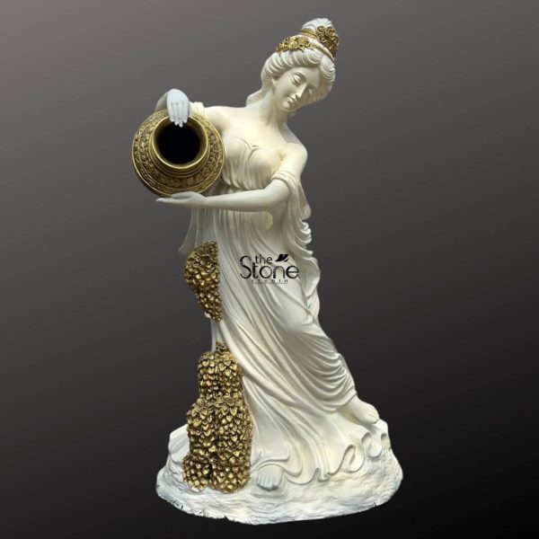 A striking 6-foot fiber Roman Lady Holding Urn fountain statue stands elegantly against a neutral dark background. The statue is primarily white depicting a graceful female figure draped in flowing classical attire. She wears a golden crown or intricate headpiece and her hair is styled in an elaborate updo. Her head is tilted downwards and her gaze is directed towards the golden urn she holds delicately in her hands positioned to appear as if pouring water. The urn itself is a rich ornate gold providing a stark contrast to the white of the statue. Cascading down the front of her gown from her waist to her feet are decorative golden elements resembling clusters of leaves or grapes suggesting a theme of abundance or nature often associated with classical figures. The base of the statue is also white textured to simulate natural ground or rock. This detailed image highlights the artistic craftsmanship and classical inspiration of the fiber Roman lady fountain statue a key subject of the accompanying article.