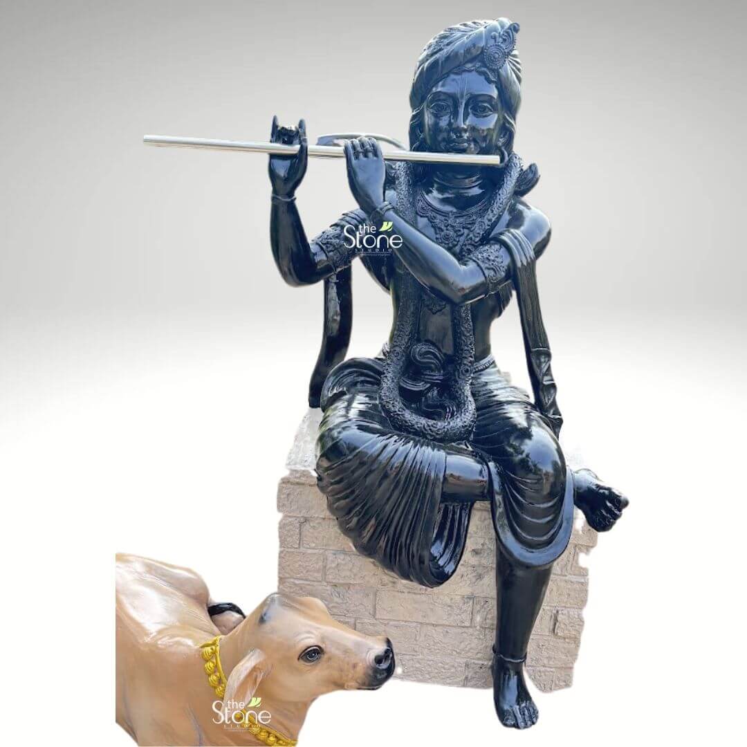 Lord Krishna with Cow Fibre Statue 4ft 5 An exquisitely crafted high-gloss black fibre statue of Shri Krishna Murli Dhar, depicted in a serene seated posture atop a light grey, stone-textured pedestal. The deity is shown in his iconic flute-playing form, holding a sleek silver-toned flute to his lips with a peaceful and meditative expression. He is adorned with a highly detailed turban, layered necklaces, and armlets that glisten against the polished black finish of the fibre sculpture. Resting faithfully at the base is a lifelike, light-brown sacred cow decorated with a vibrant yellow beaded garland, symbolizing the divine bond of the celestial cowherd. This durable and lightweight spiritual icon serves as a stunning centerpiece for a home mandir, luxury foyer, or as outdoor garden decor, bringing an atmosphere of tranquility and devotion to any setting.