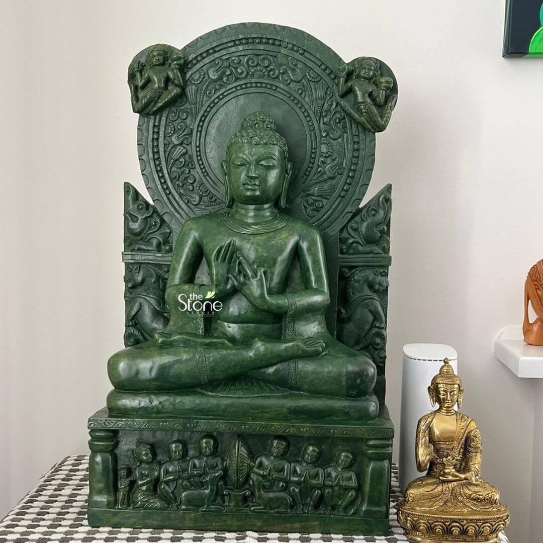 Meditation Buddha Statue 2ft 3 An exquisite green stone Buddha statue serves as a serene spiritual centerpiece in a contemporary home in Melbourne, Australia. This masterfully hand-carved stone sculpture depicts Lord Buddha in the Dharmachakra Mudra (the gesture of teaching), seated in a meditative lotus posture atop a rectangular pedestal that features detailed relief carvings of devotees. The idol is magnificently framed by an ornate circular backplate (Prabhavali) embellished with intricate traditional motifs and celestial figures. Complementing the large green statue is a smaller, gold-finished Buddha idol seated on an intricate lotus base in the Abhaya Mudra, symbolizing protection and peace. Both sacred pieces are thoughtfully arranged on a stylish black-and-white checkered cloth against a minimalist white wall, creating a sophisticated and tranquil meditation corner. This setting highlights how traditional stone statues can be harmoniously integrated into modern Australian interior design, bringing a sense of ancient heritage and spiritual grounding to urban living spaces.