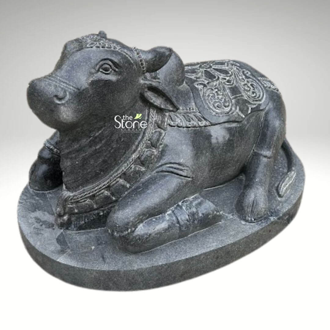 2ft Nandi Bull Stone Idol 9 An exquisitely hand-carved 2-foot tall black stone Nandi bull statue masterfully designed in a traditional temple style to serve as a profound spiritual centerpiece for a home mandir or temple sanctuary. This black stone Nandi idol depicts the sacred bull the loyal vehicle (vahana) of Lord Shiva in a serene and stable recumbent posture atop a solid integrated oval pedestal. The sculpture showcases exceptional craftsmanship with highly intricate detailing. Nandi is adorned with traditional temple ornaments including multiple layers of carved bell necklaces around its sturdy neck and a decorative headpiece positioned between its ears. A richly detailed caparison featuring a rhythmic floral relief pattern and fine borders is draped across its back and prominent hump reflecting the grandeur of heritage Indian stone masonry. The deep charcoal hue and the naturally grainy texture of the stone emphasize the artisan's attention to detail giving the idol a timeless, rustic appeal. This durable and weather-resistant 2ft Nandi sculpture is ideal for creating an atmosphere of devotion and strength in any sacred space whether positioned at the threshold of a grand temple or within a personal prayer room, designed to invite divine protection and peace into the environment.