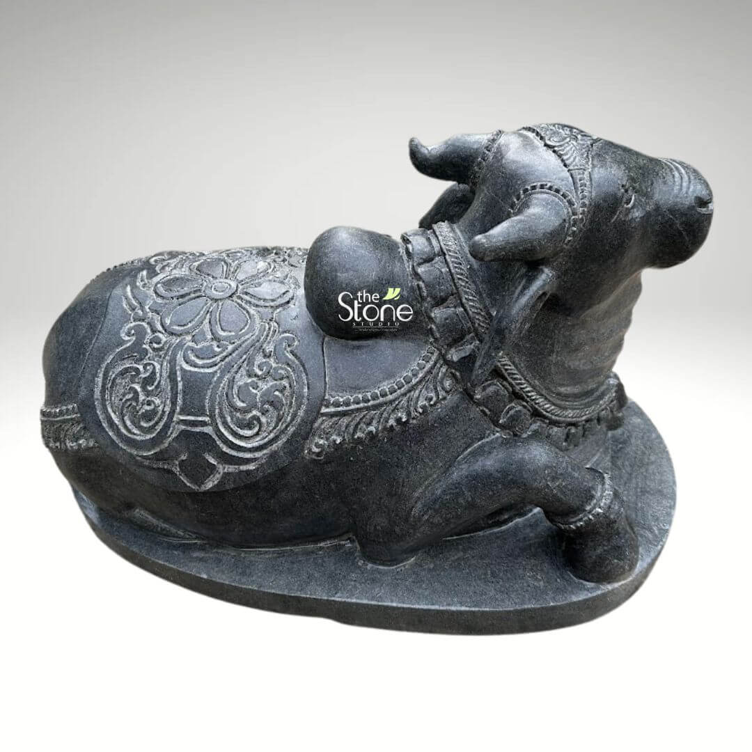 2ft Nandi Bull Stone Idol 8 An exquisitely hand-carved 2-foot tall Nandi idol masterfully sculpted from premium natural black stone with a refined matte rustic finish. Shown from an overhead three-quarter rear perspective, the sculpture highlights the exceptional 360-degree craftsmanship required for a traditional Shiva temple centerpiece. The back of the sacred bull is adorned with a highly detailed caparison featuring a large central floral mandala and intricate geometric relief patterns etched into the stone. Lord Nandi is depicted in a stable and serene recumbent posture atop a solid integrated oval pedestal. Masterful artisan detailing is further visible in the layered bell necklaces around its neck and the ornate headpiece positioned between its ears. This durable and weather-resistant black stone Nandi statue perfectly captures the divine essence of Lord Shiva’s loyal vehicle symbolizing strength, patience, and unwavering devotion. Its timeless aesthetic and significant size make it an ideal spiritual icon for a home mandir, a grand temple entrance, or an outdoor meditation sanctuary, designed to invite auspicious energy into any sacred environment.