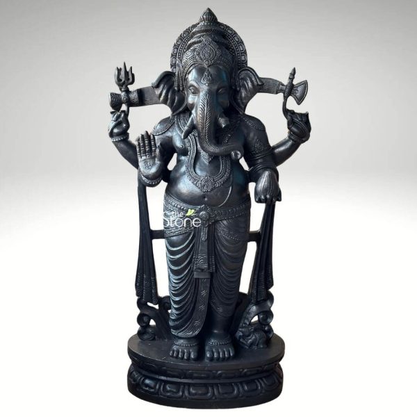 An exquisitely hand-carved Vastu Ganesha standing statue in black stone, capturing the Lord of Beginnings in a powerful and auspicious "Sthanaka" (standing) pose. Masterfully sculpted from high-quality natural black stone, the idol features a deep charcoal hue with a polished, smooth finish that highlights the intricate details of the craftsmanship. Lord Ganesha is depicted with four arms, holding traditional spiritual symbols: his upper right hand holds a trishula (trident), and his upper left hand holds a parashu (axe), representing the removal of obstacles and the cutting of worldly attachments. His lower right hand is raised in the Abhaya Mudra, the gesture of fearlessness and blessing, while his lower left hand rests gracefully on a long mace (Gada). The deity’s elephant head is adorned with a towering, multi-tiered crown (mukut) featuring fine filigree-like patterns. He wears layered necklaces, armlets, and a waist belt (kamarband), with his traditional dhoti showcasing realistic, flowing fabric folds. Standing firmly on a circular lotus pedestal embellished with relief carvings, the statue also includes a small figure of his loyal vehicle, the mouse (Mooshika), at his feet. This black stone Ganesha sculpture serves as a perfect spiritual centerpiece for a home entrance, office, or mandir, designed to invite positive energy, protection, and prosperity into any living space according to Vastu principles.