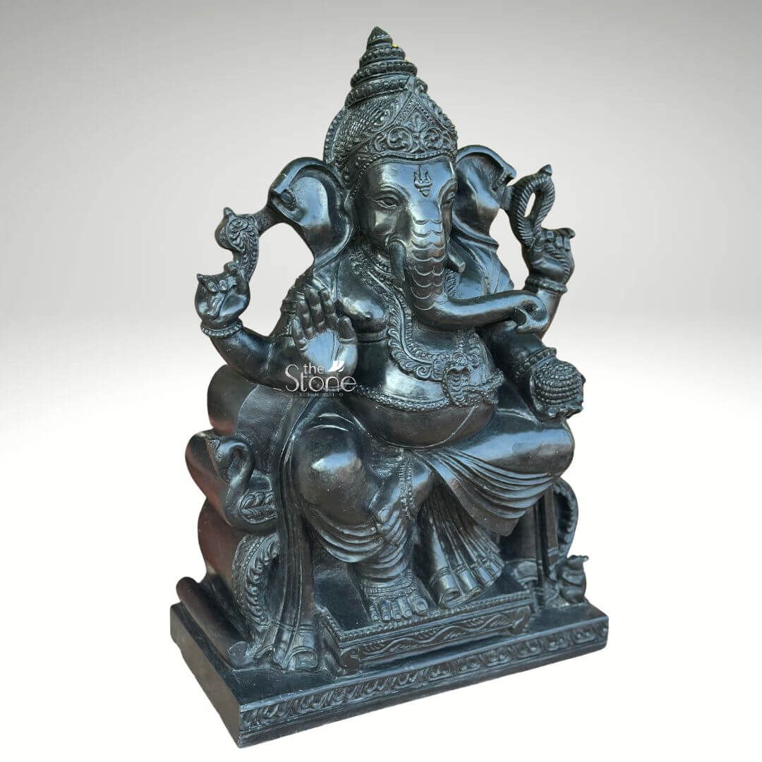 Black Stone Ganesha Idol for Entrance 2ft 2 An exquisitely hand-carved 2-foot tall Lord Ganesha statue made of premium natural black stone, shown from a three-quarter perspective to highlight its depth and masterful craftsmanship. This spiritual idol depicts Ganesha in a stable and serene seated posture atop an ornate rectangular pedestal, making it an ideal centerpiece for a home temple or sacred prayer space. The deity is portrayed with four arms carrying traditional Vedic symbols: his upper hands hold a pasha (noose) and ankusha (goad) to guide and remove obstacles, while his lower right hand is raised in the Abhaya Mudra, the universal gesture of protection and blessing. His trunk curves gracefully toward a bowl of modaks held in his lower left hand, symbolizing the sweetness of spiritual attainment. The sculpture features incredible detailing, from the towering, tiered mukut (crown) and large expressive ears to the layered necklaces, armlets, and realistic fabric folds of his traditional attire. The refined matte charcoal finish of the blackstone Ganesha sculpture highlights the heritage of Indian stone masonry, inviting an atmosphere of peace, prosperity, and divine stability into any contemporary or traditional home environment.