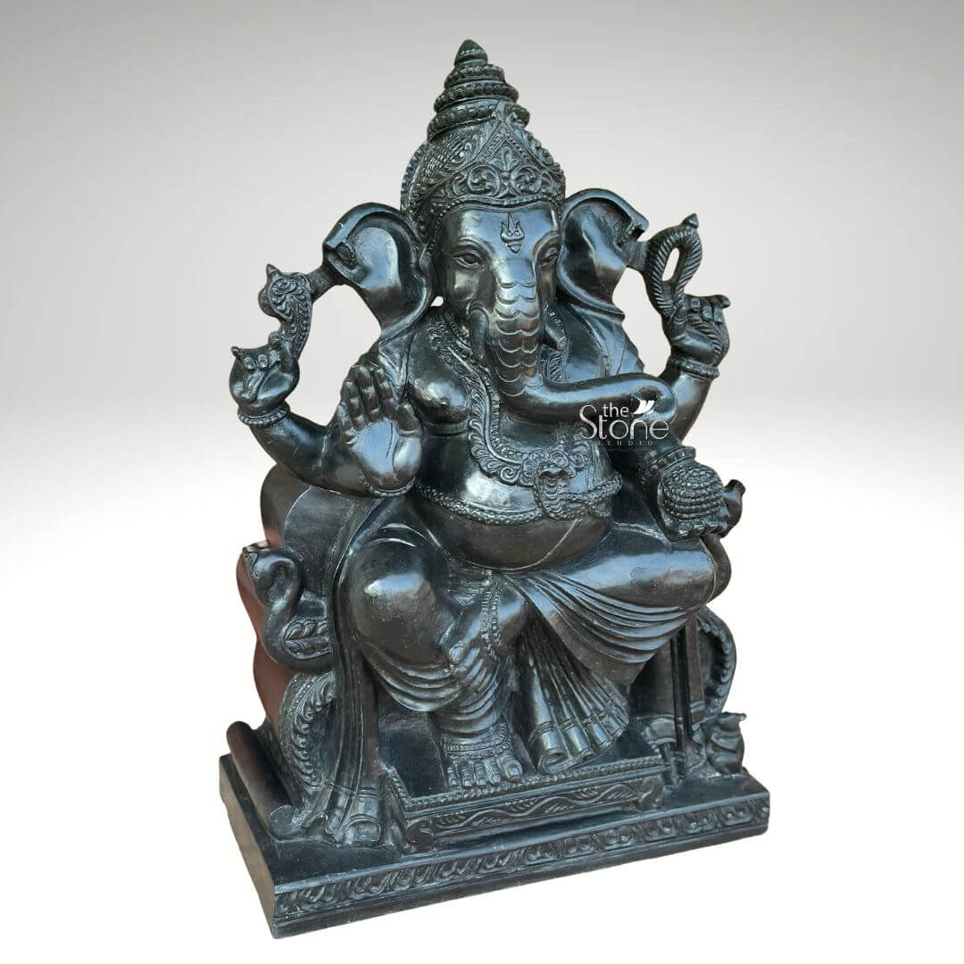 Black Stone Ganesha Idol for Entrance 2ft 4 An expertly hand-carved 2-foot tall seated Lord Ganesha statue masterfully sculpted from premium natural black stone. This spiritual masterpiece, shown from a three-quarter side angle to emphasize its depth and volume, captures the deity in a stable and serene seated posture atop a rectangular pedestal embellished with intricate relief carvings. Lord Ganesha is portrayed with four arms carrying sacred Vedic symbols: his upper hands hold traditional implements used to remove obstacles and guide devotees, while his lower right hand is raised in the Abhaya Mudra, the universal gesture of protection and blessing. His trunk curves gracefully toward a bowl of modaks (traditional sweets) held in his lower left hand, symbolizing the rewards of a spiritual life. The craftsmanship is highly detailed, featuring a towering, tiered Mukut (crown), large expressive fan-like ears, and multiple layers of ornate necklaces and armlets. The deity's traditional dhoti is carved with realistic, fluid fabric folds that add a sense of elegance to the solid stone. The refined matte charcoal finish of the blackstone Ganesha idol highlights the heritage of traditional Indian stone masonry, making it an ideal choice for a home mandir, entrance foyer, or meditation space to invite peace and prosperity.