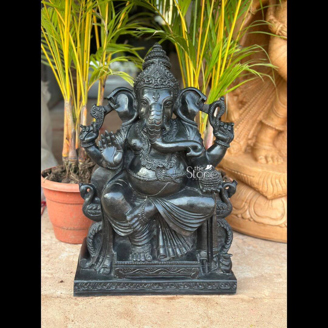 Black Stone Ganesha Idol for Entrance 2ft 3 An exquisitely hand-carved 2-foot tall Ganesh Ji murti made from premium natural black stone, serving as a majestic spiritual centerpiece for a home or garden. The sculpture depicts Lord Ganesha in a stable and auspicious seated posture atop a rectangular pedestal embellished with traditional relief carvings. The deity is shown with four arms holding sacred symbols: his upper hands carry the pasha (noose) and ankusha (goad) to remove obstacles, while his lower right hand is raised in the Abhaya Mudra, the universal gesture of protection and blessing. His trunk curves gracefully toward a bowl of modaks (sweets) held in his lower left hand, representing the sweetness of spiritual fulfillment. The craftsmanship is highly detailed, featuring a towering tiered mukut (crown), large expressive fan-like ears, and layered necklaces carved directly into the stone. Photographed in an outdoor setting, the refined matte charcoal finish of the black stone Ganesha statue contrasts beautifully with the vibrant yellow-green fronds of a palm plant and terracotta pots in the background. This large-scale natural stone Ganesha idol perfectly illustrates how traditional Indian stone masonry can be integrated into modern landscapes or home temples to invite peace and prosperity.