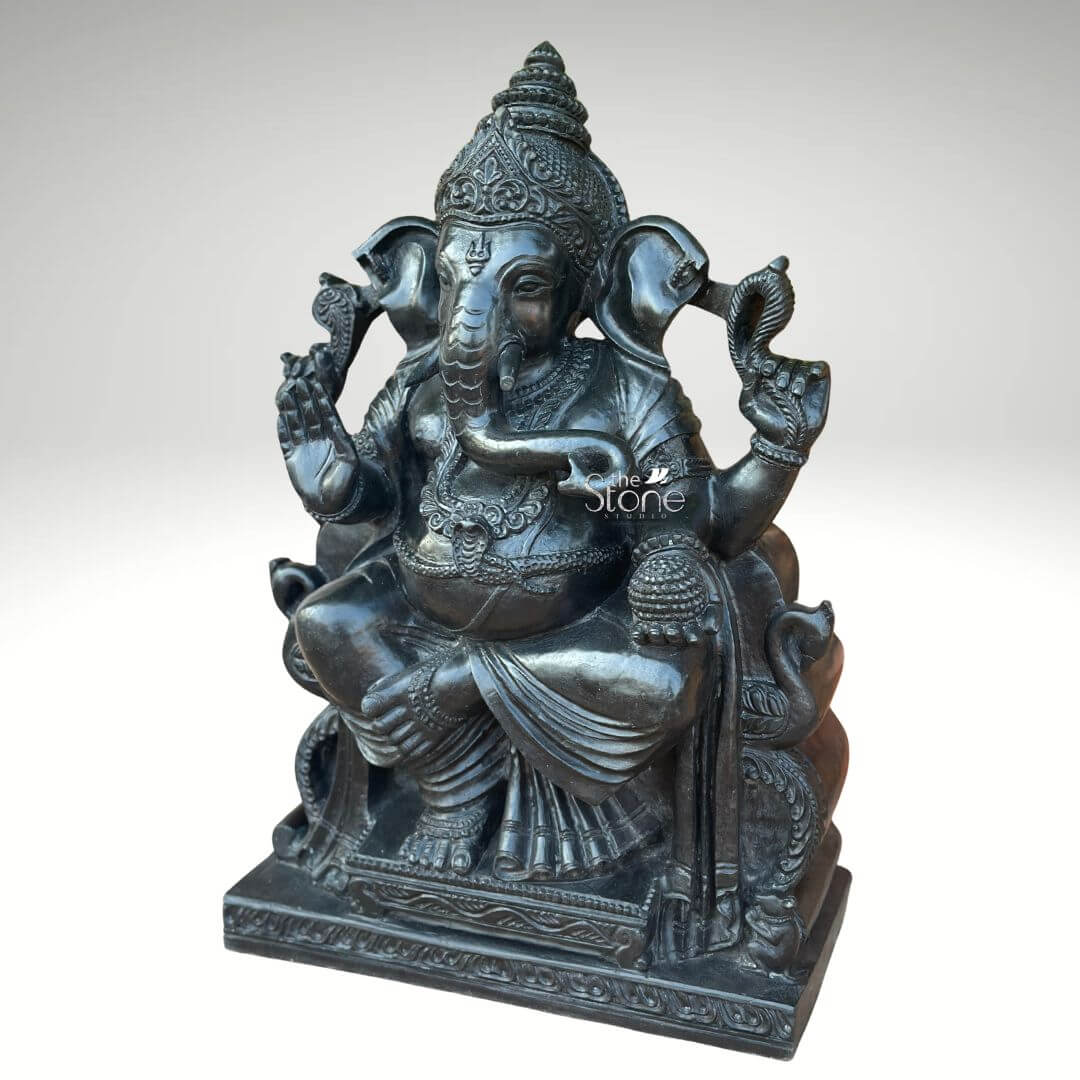 Black Stone Ganesha Idol for Entrance 2ft 5 An exquisitely handcrafted 2 ft Ganpati Bappa idol masterfully carved from natural black stone, serving as a majestic spiritual centerpiece for home decor. Shown from a three-quarter perspective to highlight its impressive depth and volume, this 2-foot Ganesha statue depicts the deity in a stable and auspicious seated posture. The sculpture features intricate detailing on the deity's towering mukut (crown), large expressive ears, and gracefully curved trunk. Lord Ganesha is portrayed with four arms holding sacred Vedic symbols, including a bowl of modaks (traditional sweets) and traditional implements to remove obstacles. His lower right hand is raised in the Abhaya Mudra, the universal gesture of protection and blessing. The idol is further adorned with layered necklaces, armlets, and a sacred snake thread (yajnopavita) draped across his torso. Every fold of the traditional dhoti and the ornate patterns on the multi-tiered pedestal are meticulously rendered, showcasing the heritage of traditional Indian stone masonry. With its refined matte charcoal finish, this durable and timeless black stone Ganesha sculpture is an ideal addition to invite peace, prosperity, and divine energy into a home mandir, entrance foyer, or meditation space.