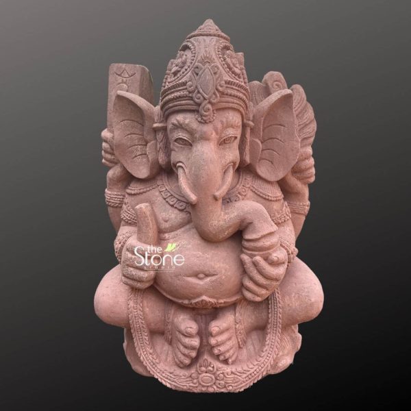 This image features a charming 2-foot tall Bali Ganesha Murti expertly carved from sandstone presented against a dark, neutral background. Lord Ganesha is depicted seated in a relaxed and slightly squatting posture, which is characteristic of some Balinese or traditional Indian artistic styles. Ganesha has a prominent round belly symbolizing prosperity. His trunk curves gracefully to his left and he holds his iconic broken tusk in one of his lower right hands. His other hands hold traditional attributes, though some details are softly rendered due to the sandstone texture. He is adorned with sculpted jewelry, including necklaces, armlets and a sacred thread (yajnopavita), all carved into the stone. His large ears expressive eyes and ornate tiered crown (mukuta) with a central jewel motif add to his divine appearance. The texture of the sandstone material is clearly visible throughout the sculpture giving it an earthy natural, and slightly rustic feel enhancing its antique appeal. The base on which Ganesha sits is also carved with decorative elements, including a floral border along the bottom. This 2ft Bali Ganesha Murti embodies both spiritual significance and the unique aesthetic of traditional craftsmanship making it an ideal piece for various settings.