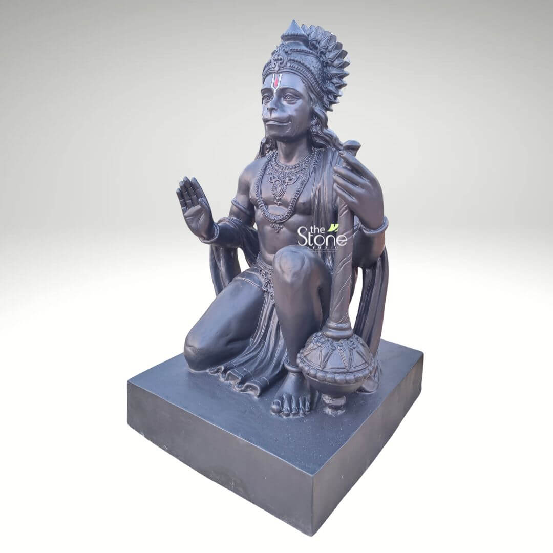 3 Feet Hanuman Ji Statue 2 A majestic 3-foot tall Lord Hanuman (Bajrangbali) idol expertly hand-crafted from high-quality fibre material, showcased in a three-quarter side view to highlight its depth and masterful detail. The sculpture features a sophisticated matte charcoal black finish that replicates the timeless aesthetic of natural stone while remaining lightweight and durable. Lord Hanuman is depicted in a powerful kneeling posture, with his right hand raised in the Abhaya Mudra, the universal gesture of protection and blessing, and his left hand firmly holding his signature Gada (mace) positioned vertically. The idol is adorned with an intricate, tiered crown (mukut), layered necklaces, and traditional armlets, featuring a prominent sacred tilak on his forehead. Resting on a solid rectangular pedestal of the same charcoal hue, this large-scale spiritual sculpture serves as a perfect centerpiece for a grand home mandir, professional office foyer, or as auspicious outdoor garden decor. This 36-inch fibre Hanuman statue is designed to withstand environmental elements while inviting strength, peace, and divine protection into any modern or traditional living space.
