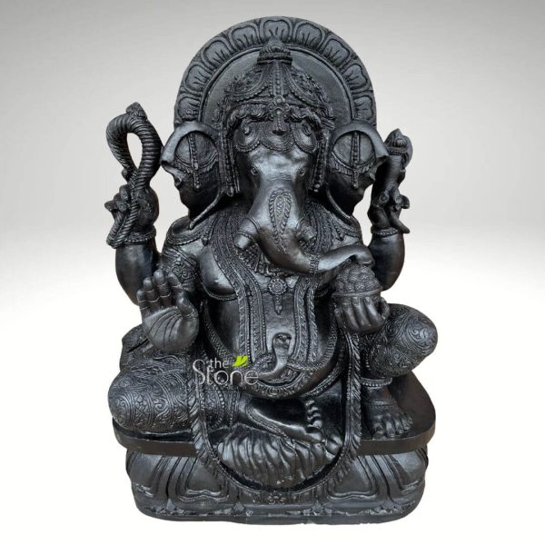 A majestic 3-foot tall Ganapati stone idol, masterfully hand-carved from premium natural black stone, serves as a profound spiritual centerpiece for a home mandir or meditation space. The sculpture depicts Lord Ganesha in a stable and auspicious seated posture, radiating a sense of calm and divine protection. The deity is shown with four arms, holding traditional Vedic symbols: his upper hands carry the pasha (noose) and ankusha (elephant goad) to guide and remove obstacles, while his lower left hand holds a bowl of modaks, representing the sweetness of spiritual fulfillment. His lower right hand is raised in the Abhaya Mudra, the universal gesture of fearlessness and blessing. The craftsmanship is highly detailed, featuring a towering, multi-tiered Kiritamukuta (crown), large expressive fan-like ears, and a gracefully curved trunk. The idol is adorned with intricate jewelry, including layered necklaces, armlets, and a sacred serpent thread (Yajnopavita) draped across his torso. Framing the deity is a grand, circular Prabhavali (ornate arch) embellished with rhythmic petal-like relief patterns that symbolize a divine aura. Resting on a tiered rectangular pedestal with lotus-petal carvings, this 3-foot blackstone Ganesha statue combines ancient Indian stone masonry with a refined matte finish. It is a durable and weather-resistant spiritual icon, ideal for inviting peace, prosperity, and positive energy into a contemporary or traditional home environment.