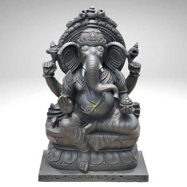 An exquisitely hand-carved 3-foot tall Ganpati Bappa idol, masterfully sculpted from premium natural blackstone to serve as a majestic spiritual centerpiece. This large-scale black stone Ganesha statue depicts the deity in an auspicious and stable seated posture atop an intricate, multi-tiered lotus pedestal. Lord Ganesha is portrayed with four arms carrying sacred Vedic symbols: his upper hands hold the Sudarshana Chakra (discus) and the Panchajanya Shankha (conch), while his lower right hand is raised in the Abhaya Mudra, the universal gesture of protection and blessing. His trunk curves gracefully toward a bowl of modaks (traditional sweets) held in his lower left hand, symbolizing the rewards of a spiritual life. The craftsmanship showcases incredible detail, from the towering, tiered Mukut (crown) and large expressive ears to the layered necklaces, armlets, and realistic fabric folds of his traditional attire. A grand circular Prabhavali (ornate arch) frames the deity’s head, representing a divine aura. At the base of the statue, the small figure of his loyal vehicle, the mouse (Mooshika), is subtly carved. The deep charcoal finish of the stone highlights the heritage of traditional Indian stone masonry, making this 36-inch Ganesha idol a perfect addition to a home mandir or grand foyer to invite peace, prosperity, and the removal of obstacles.