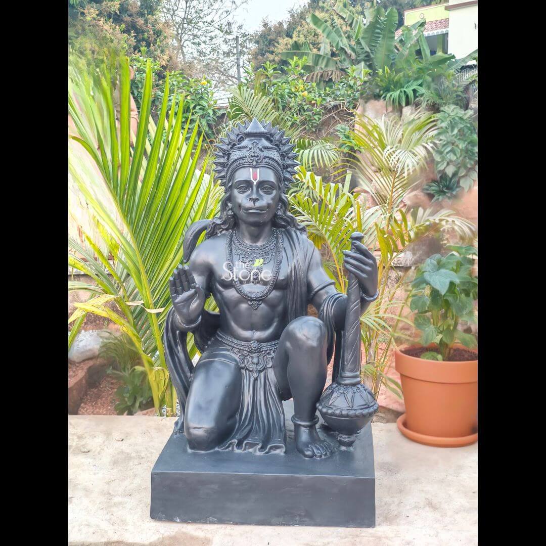 3 Feet Hanuman Ji Statue 3 A majestic 3-foot tall Lord Hanuman murti crafted from high-quality fibre material is showcased as a powerful spiritual centerpiece in a lush outdoor garden. The sculpture is finished in a sophisticated matte charcoal black, mimicking the refined look of natural stone while offering the lightweight durability of modern fibre. Lord Hanuman is depicted in a traditional kneeling devotional pose on a solid rectangular base. His right hand is raised in the Abhaya Mudra, a universal gesture of protection and blessing, while his left hand firmly holds his signature Gada (mace) resting on the ground. The idol is meticulously detailed with a towering, sunburst-style crown (mukut), layered necklaces, and armlets. His serene facial expression, prominent white tilak, and flowing hair add a lifelike and divine aura to the piece. Set against a vibrant backdrop of tropical greenery, including large palm fronds and terracotta-potted plants, this 36-inch fibre Hanuman statue illustrates how large-scale spiritual art can be integrated into residential or temple landscapes. Its weather-resistant construction makes it an ideal choice for creating a peaceful meditation sanctuary or an auspicious home entrance, inviting strength, peace, and protection into the surroundings.