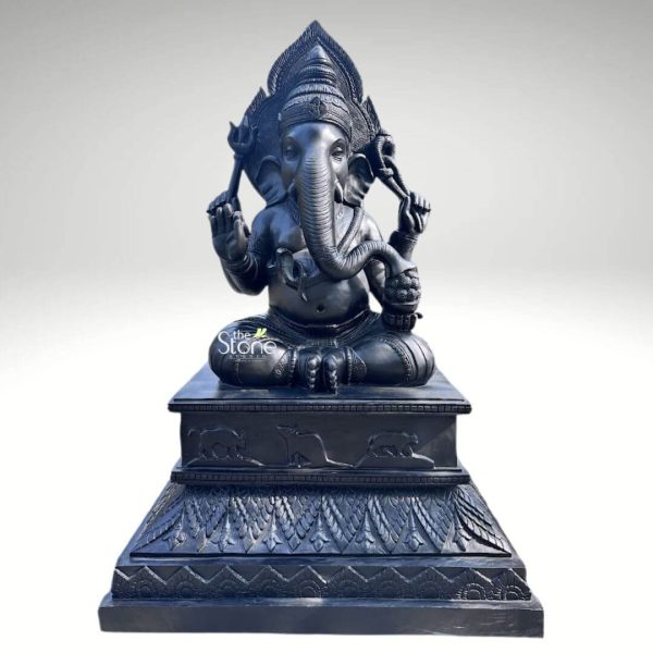 A magnificent 8-foot tall Lord Ganesha statue, masterfully hand-carved from premium natural black stone, serves as a majestic spiritual centerpiece for a temple foyer or sanctuary. This large-scale blackstone Ganesha idol is depicted in a stable seated posture atop an exceptionally wide and intricately detailed multi-tiered pedestal. Lord Ganesha is portrayed with four arms carrying sacred symbols: his upper hands hold a trident (trishula) and a noose (pasha), while his lower right hand is raised in the Abhaya Mudra, the universal gesture of protection and blessing. His trunk curves gracefully toward a bowl of modaks (traditional sweets) held in his lower left hand, representing the sweetness of spiritual fulfillment. The craftsmanship is highly detailed, featuring a towering, tiered mukut (crown), large expressive fan-like ears, and layered necklaces carved directly into the stone. The massive stepped base is adorned with relief carvings of Ganesha’s vahana, the mouse (mooshika), along with rhythmic floral and geometric patterns. The refined matte charcoal finish of the stone highlights the heritage of traditional Indian stone masonry, providing an aura of stability, permanence, and divine grandeur suitable for a prominent temple installation.