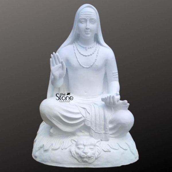 A magnificent 3-foot tall fiber statue of Lord Adi Shankaracharya, depicted in a serene seated meditative posture. This intricately hand-carved white sculpture features the great philosopher-saint with his right hand raised in the Abhaya Mudra, offering protection and blessing, while his left hand rests in his lap holding a sacred palm-leaf manuscript, symbolizing his profound contributions to Advaita Vedanta and spiritual knowledge. The statue displays a peaceful facial expression with a traditional Tripundra tilak on the forehead and is adorned with multiple layers of realistic bead necklaces. Lord Shankaracharya is shown seated upon a textured rocky base featuring a meticulously carved tiger-skin seat, a traditional iconographic element representing a realized sage. Crafted from high-quality white fiber, the idol has the elegant, polished appearance of marble while remaining lightweight and durable. This Adi Shankaracharya idol serves as a perfect spiritual centerpiece for a home mandir, meditation room, or educational library, designed to inspire an atmosphere of ancient wisdom and divine tranquility.