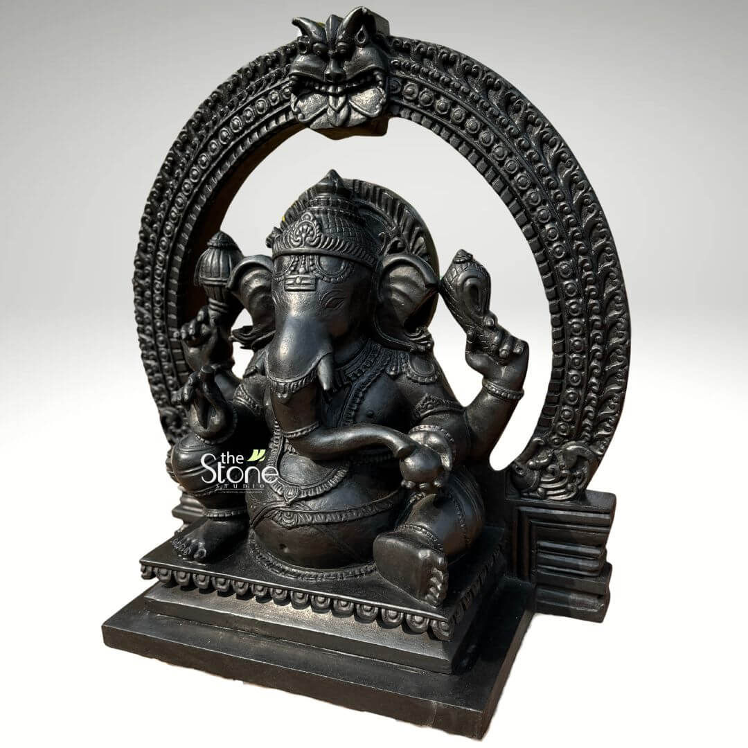 Vighnaharta Ganesh Idol 2ft 2 An exquisitely hand-carved 24-inch black stone Ganesh idol with a decorative arch, masterfully crafted from high-quality fibre with a refined black stone finish. This three-quarter view highlights the depth and traditional craftsmanship of the 2-foot spiritual sculpture, making it a perfect focal point for a home mandir, office, or meditation space. The idol depicts Lord Ganesha in a stable and serene seated meditative pose. He is framed by a grand, circular Prabhavali (ornate arch) featuring a protective Kirtimukha face at the peak and a repetitive textured pattern along its circumference. The deity is shown with four arms holding sacred symbols, including a Gada (mace) and a Shankha (conch shell), while his large expressive ears and curved trunk symbolize wisdom and deep listening. Every detail, from the tiered mukut (crown) to the layered necklaces and the fluid fabric folds of his traditional attire, is rendered with exceptional precision. The statue rests on a solid, multi-tiered rectangular pedestal embellished with a lotus-petal relief border. This durable and lightweight fibre Ganesha statue offers the majestic appearance of natural blackstone, designed to invite peace, stability, and auspicious energy into any contemporary or traditional living environment.