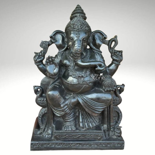 A majestic 2-foot tall Lord Ganesha idol meticulously hand-carved from solid natural black stone, designed to serve as a powerful spiritual centerpiece for a home entrance. The statue captures Ganesha in a stable and serene seated pose atop an ornate rectangular pedestal. The deity is depicted with four arms, holding traditional sacred symbols—the pasha (noose) and ankusha (goad)—to guide and remove obstacles, while his lower right hand is raised in the Abhaya Mudra, a universal gesture of protection and blessing. His lower left hand holds a bowl of modaks, representing the rewards of spiritual pursuit. The sculpture features a refined matte-charcoal finish that highlights the intricate artisan details, from the towering tiered crown (mukut) and expressive fan-like ears to the layered necklaces and the realistic, fluid folds of his traditional dhoti. This durable blackstone Ganesha sculpture embodies stability and auspicious energy, making it an ideal choice for an entrance or foyer to welcome guests and invite prosperity, peace, and protection into the household.