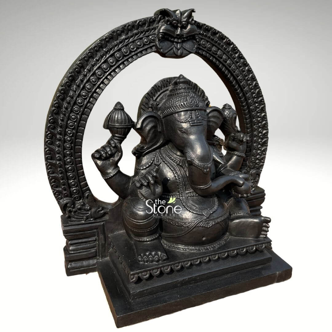 Vighnaharta Ganesh Idol 2ft 4 An intricately hand-carved 24-inch black stone Ganesha statue with an arch, masterfully crafted from high-quality fibre with a refined blackstone finish. This 2-foot spiritual idol depicts Lord Ganesha in a stable and serene seated posture, making it a perfect centerpiece for a home mandir, office entrance, or meditation space. The sculpture features the deity framed by a grand, circular Prabhavali (ornate arch), which is embellished with detailed repetitive patterns and a protective Kirtimukha face at the peak, adding a sense of ancient temple grandeur. Lord Ganesha is shown with four arms holding sacred symbols, including a Gada (mace) and a Shankha (conch shell), while his large expressive ears and curved trunk represent deep wisdom and the removal of obstacles. Every element, from the tiered mukut (crown) to the layered necklaces and the fluid fabric folds of his traditional attire, is rendered with exceptional precision. The statue rests on a solid, multi-tiered rectangular pedestal featuring a lotus-petal relief border. This durable and lightweight fibre Ganesha statue offers the timeless aesthetic of natural black stone, designed to invite peace, stability, and auspicious energy into any contemporary or traditional living environment.
