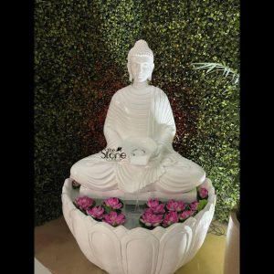 A serene and majestic 5-foot tall white fiber Lord Buddha fountain, designed as a tranquil spiritual centerpiece for a modern home or garden in Delhi. The sculpture depicts Buddha in a peaceful meditative seated posture, featuring a smooth, polished finish that captures the elegant appearance of fine white stone or marble. Water cascades gently from a sacred bowl held in the deity's hands, creating the soothing sound of falling water. The statue is positioned atop a wide, beautifully carved lotus-shaped water basin filled with several vibrant pink lotus blossoms. The entire Buddha fountain is set against a lush, dark green vertical garden wall, which provides a natural backdrop that emphasizes the statue's luminous white presence. This large-scale fiber water feature perfectly illustrates how a Buddha fountain in Delhi can transform an urban space—be it a residential backyard, luxury terrace, or commercial foyer—into a peaceful Zen retreat, blending traditional spiritual iconography with modern, durable, and lightweight materials.