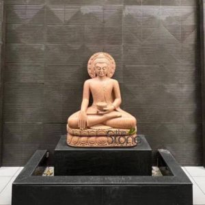 An exquisitely hand-carved stone Lord Buddha statue serves as a serene spiritual centerpiece in a luxury residential setting in Aamby Valley, Lonavala. The sculpture, crafted from warm-toned natural sandstone, depicts Buddha in a meditative seated pose atop an intricately detailed lotus pedestal. He is shown with one hand in the Bhumisparsha mudra (earth-touching gesture) and the other holding a sacred bowl, with a beautifully ornate circular halo (Prabhavali) framing his head. The idol is elegantly positioned atop a tiered black granite platform, which integrates into a minimalist modern landscape design. The background features a contemporary dark grey stone wall with a sophisticated hatched geometric pattern, creating a striking contrast that highlights the deity's presence. This setting perfectly exemplifies how high-quality stone Buddha statues can enhance the tranquility and aesthetic appeal of upscale villas and landscapes in Lonavala, inviting an aura of peace and mindfulness into the environment.