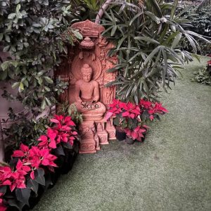 A majestic 4-foot tall Lord Buddha meditation statue hand-carved from natural brown sandstone, serving as a serene spiritual centerpiece in a lushly landscaped Gurgaon, Haryana garden. The sculpture depicts the Buddha in a tranquil seated Padmasana posture with hands resting in the Dhyana Mudra, signifying deep inner peace and mindfulness. The idol is framed by an exceptionally detailed stone stele (back panel) that features traditional Indian iconography, including a circular halo and a protective stone canopy. The statue is artfully nestled between vibrant red Poinsettia flowers and lush tropical green palms, positioned on a manicured green lawn. The warm, earthy tones and rustic texture of the brown sandstone contrast beautifully with the vibrant garden foliage, illustrating how a large-scale Buddha idol can be integrated into residential or commercial landscapes in Gurgaon to create a peaceful Zen sanctuary for reflection and devotion.