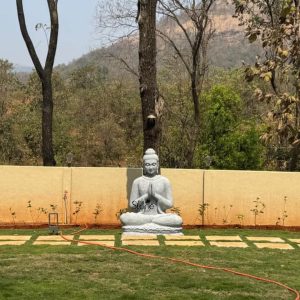 A serene and majestic grey-textured Buddha statue sits in a cross-legged meditative posture with hands joined in the Anjali Mudra (namaste gesture), serving as a peaceful spiritual centerpiece in a lush garden in Karjat, near Mumbai. The large-scale outdoor sculpture, crafted with a natural stone-like finish, is positioned on a vibrant green manicured lawn accented by a geometric stone tile pathway. In the background, a minimalist beige boundary wall separates the garden from a natural forested area and the prominent, sun-lit rocky hills typical of the Maharashtra landscape. This setting perfectly illustrates how a stone Buddha idol can enhance the Zen-like atmosphere of a residential or resort outdoor space, providing a tranquil focal point for meditation and mindfulness in the scenic outskirts of Mumbai.