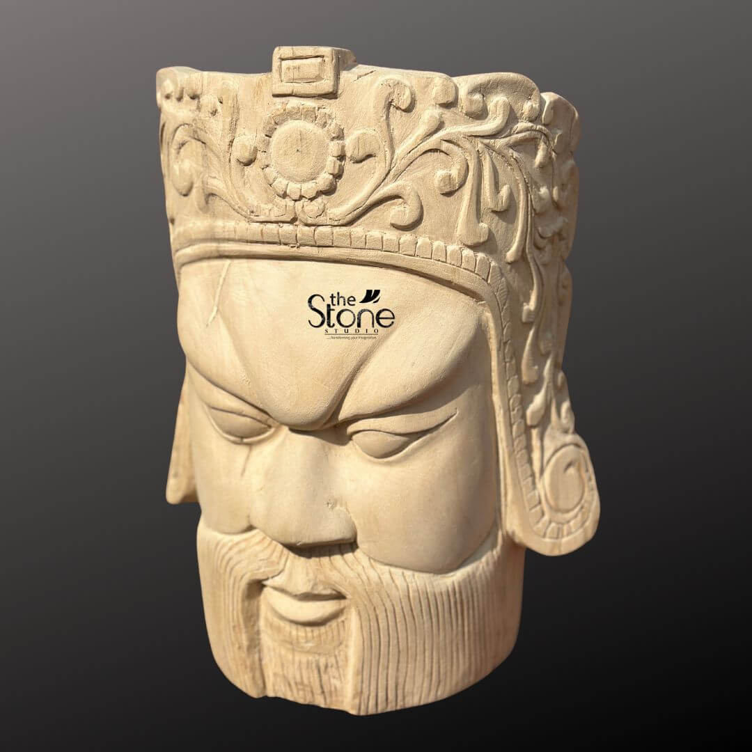 Royal Head Sculpture 12" 2 This image showcases a striking 12-inch Decorative Wooden Face Bust, meticulously carved from light-colored wood. The sculpture depicts a male head, possibly a warrior or a deity, with a stern and commanding expression, presented against a dark, neutral background. The face features strong, angular characteristics: deeply furrowed brows, intense, narrow eyes that gaze downward, a straight nose, and a firmly closed mouth adorned with a sculpted beard and mustache. The expression conveys a sense of power and gravity, making it a compelling decorative piece. Crowning the head is an elaborate headdress or helmet, richly detailed with intricate floral patterns, swirling motifs, and a central circular emblem, possibly a sun or a stylized flower. The precision of the carving highlights the skill involved, with every element meticulously shaped from the wood. The natural grain and subtle imperfections of the wood are visible throughout the bust, especially on the face and beard, adding to its organic texture and handcrafted appeal. This wooden head sculpture serves as an impactful and artistic focal point, blending historical or mythological aesthetics with natural material.