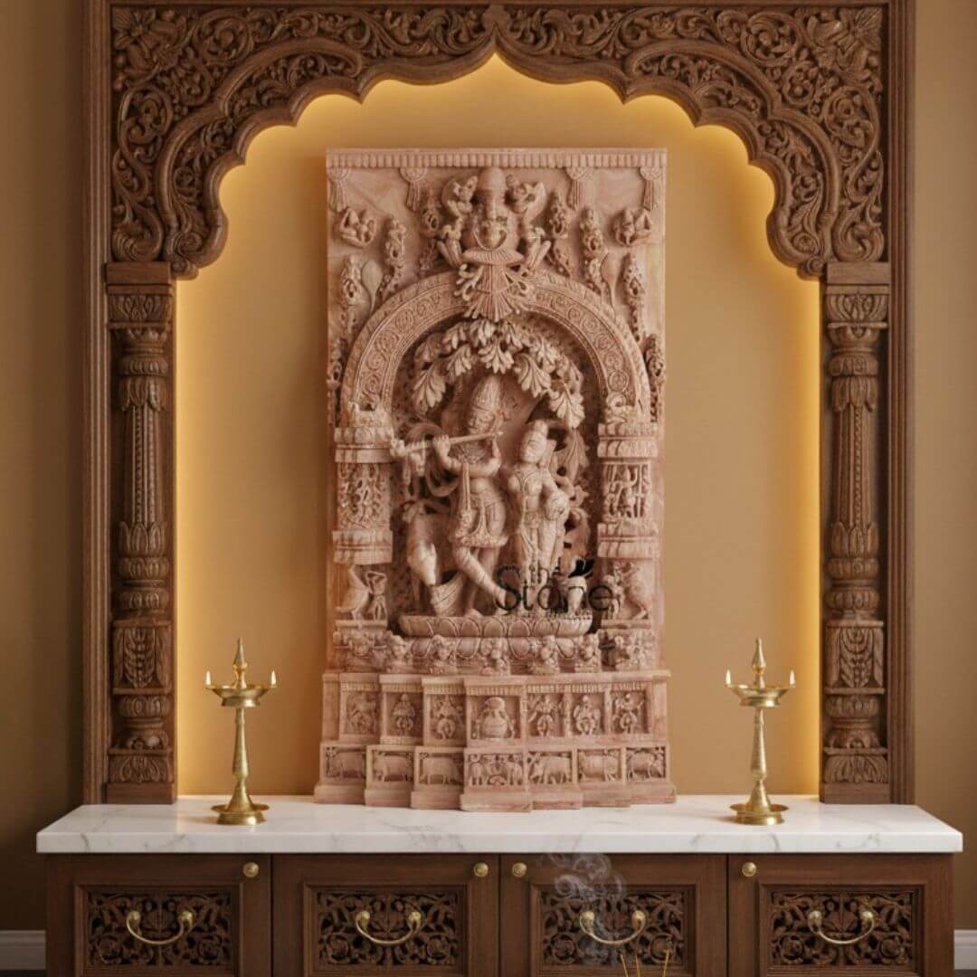 Pink Stone Radha Krishna Murti for Home 4ft 3 A stunning 4-foot tall pink stone Radha Krishna sculpture is elegantly displayed within a warm, illuminated wooden archway, creating a focal point for a home altar or sacred space. The exquisite stone carving features Lord Krishna playing his flute under a meticulously detailed tree, with his beloved Radha standing beside him, their forms rendered with grace and devotion. The sculpture is rich with intricate details: Krishna's peacock feather crown, their delicate ornaments, and the flowing drapery of their attire are all beautifully carved from the pink stone. Above the central figures, the archway of the stone panel itself is adorned with celestial beings and additional divine figures, while the sides feature smaller sculptural elements and niches, enriching the narrative. The base of the stone sculpture showcases a series of finely carved friezes and smaller scenes, adding depth and storytelling to the entire piece. The wooden archway framing the sculpture is also intricately carved with traditional Indian motifs, its warm brown tones complementing the pink stone. Soft, warm backlighting behind the arch illuminates the sculpture, highlighting its textures and casting a gentle glow on the surrounding wall. Below the arch and the sculpture, a white marble countertop rests on a dark wooden cabinet with ornately carved doors and brass handles. Two tall, slender brass oil lamps (diyas) flank the sculpture on the counter, with a subtle wisp of smoke rising from an incense stick to the right, signifying an active place of worship. This Exquisite Pink Stone Radha Krishna Sculpture for Home evokes a sense of peace and divine presence, perfect for devotional practices.