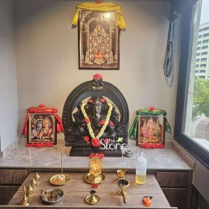 This image displays a beautifully arranged home altar featuring a 2-foot Ganapathi Idol for Home crafted from black stone, prominently displayed in a modern interior, possibly in Singapore. The central feature is the majestic black stone Lord Ganesha statue, seated in a traditional posture and adorned with fresh floral garlands of white flowers and vibrant red roses. Additional red roses are placed on his crown, ears, and hands, as well as scattered on the base. The Ganesha statue is framed by an impressive, intricately carved black stone archway which features dense traditional scrollwork and a Kirtimukha at the top. The entire Ganesha figure sits on a dark, patterned marble base. On either side of the Ganesha idol are two framed devotional pictures: one of Lord Shiva and Parvati, and another of Lord Vishnu and Lakshmi (draped in green cloth), both adorned with red roses at their bases. Above the Ganesha, a larger framed picture of a South Indian deity is hung, draped with a yellow cloth. The altar surface is a sleek, dark wood with a light-colored countertop, covered with various puja items. These include multiple brass oil lamps (diyas), incense holders (one with burning incense), small golden statues (including another Ganesha), bowls of offerings, and a plastic bottle of oil. The setup is against a neutral wall with a large window to the right, offering a view of outdoor greenery and buildings, creating an interesting blend of spiritual devotion and urban living. This arrangement highlights the Ganapathi Idol for Home as a central figure of worship and beauty.