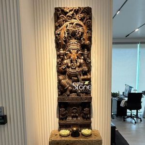 This image features a magnificent 5-foot tall "Ganesha Statue," specifically a carved wooden wall panel, prominently displayed indoors, possibly in an office or home setting in the Philippines. The panel is crafted from country wood and showcases Lord Ganesha as the central, seated figure, surrounded by an intricate tree motif and elaborate decorative carvings. Lord Ganesha is depicted in a traditional, relaxed sitting posture, with his multiple arms holding his characteristic attributes. He is richly adorned with finely sculpted jewelry, a crown, and an ornate dhoti, all rendered with deep, precise cuts that give the wood carving remarkable texture and three-dimensionality. His trunk curves gracefully to his left. Above Ganesha, an elaborate tree motif forms a magnificent canopy, with countless branches and leaves carved in high relief, creating a sense of natural grandeur. The entire background of the panel, especially the upper section and sides, is densely filled with intricate floral patterns, scrollwork, and possibly smaller divine or animal figures, highlighting the meticulous craftsmanship. The wooden Ganesha panel is mounted against a light-colored, vertically ribbed wall. It stands on a rustic stone or concrete pedestal, on top of which a small oil lamp (diya) with a flickering flame is placed, flanked by two small bowls holding white flower petals or other offerings, adding a spiritual ambiance. In the background, a modern interior space with office furniture and a large window is visible, creating an interesting contrast between traditional art and contemporary living. This wood Ganesha wall panel serves as a grand and spiritually significant focal point, bringing cultural richness to the space.