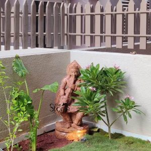 This image features a charming 3-foot tall Garden Statue,specifically a brown sandstone Devadasi figure, set in a cozy outdoor corner garden, likely in Bengaluru. The statue, carved from reddish-brown sandstone depicts a female figure in a traditional pose, seemingly playing a musical instrument held to her lips. The Devadasi figure is adorned with sculpted jewelry, a detailed headdress, and flowing garments, all carved with a rustic yet elegant finish that highlights the natural texture of the sandstone. The statue stands on a round, integrated pedestal. Importantly, there's a warm glow emanating from beneath the statue's base, suggesting integrated lighting that illuminates the figure and its immediate surroundings, creating an enchanting effect, especially at night. The statue is positioned in a small garden bed with a mix of lush greenery. To its left, a large-leafed plant grows tall, while to its right, two smaller flowering plants with delicate pink blooms add a touch of softness and color. The ground around the statue is a mix of reddish soil and green grass. The garden corner is defined by light-colored walls, and above them, a neat light brown wooden picket fence provides a charming boundary. This brown sandstone Devadasi statue serves as a beautiful and culturally rich focal point, enhancing the aesthetic of any garden space.
