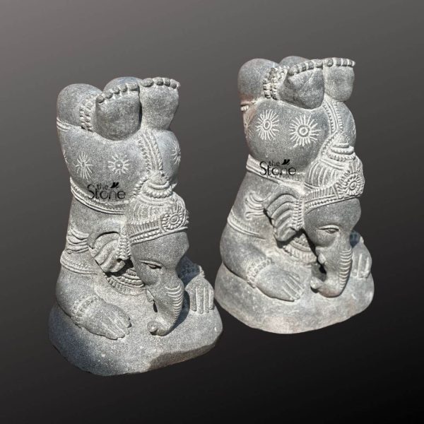 This image displays a pair of identical 16 inch Grey Stone Yoga Ganesha Statues, carved from blackstone with a distinctive textured, almost speckled appearance. Both statues depict Lord Ganesha in a unique, contemplative Yoga posture, appearing to lean forward with his trunk resting gently on a mound, suggesting deep meditation or introspection. Ganesha's large ears are visible, and his trunk curves gracefully. He is adorned with carved details that mimic traditional jewelry and sacred markings, including sunburst patterns on his body and intricate designs on his crown and forehead. His hands are positioned in a way that suggests a meditative mudra or supporting his posture, resting on his lap or the ground in front of him. The blackstone material gives the statues a robust and earthy feel, with natural variations in tone that add character. The rough, unfinished base of each statue blends naturally with the dark background, highlighting the sculptural forms. The repetitive patterns carved into his body, such as lines and dots, emphasize the artistic style. These statues, with their unique depiction of Ganesha in a yogic state, combine spiritual symbolism with artistic elegance, making them suitable for spaces dedicated to peace and mindfulness.