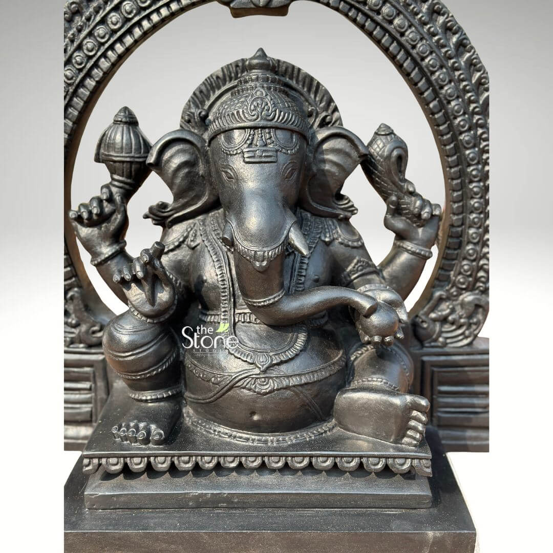 Vighnaharta Ganesh Idol 2ft 5 A highly detailed 24-inch Ganesha statue with an arch, masterfully crafted from durable fibre with a sophisticated black granite finish. This 2-foot spiritual masterpiece captures the deity in a stable, seated meditative pose, designed to replicate the majestic and heavy look of natural blackstone while remaining lightweight and easy to maintain. The sculpture features Lord Ganesha framed by a grand, circular Prabhavali (ornate arch), which is embellished with intricate repetitive patterns and a protective Kirtimukha face at its peak, evoking the grandeur of ancient Indian temples. The deity is shown with four arms holding sacred symbols, including a Gada (mace) and a Shankha (conch shell), representing divine power and the removal of obstacles. Every element of this hand-carved black stone Ganesha idol is rendered with precision, from the towering tiered crown (mukut) and layered necklaces to the realistic, fluid folds of his traditional attire. The statue rests on a solid, multi-tiered rectangular pedestal featuring a delicate relief border. This fibre Ganesha idol serves as a profound spiritual focal point, perfect for a home mandir, office entrance, or meditation space, inviting peace, stability, and auspicious energy into any living environment.