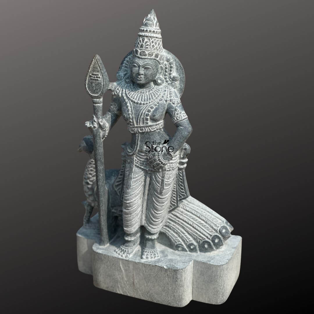 Lord Skanda Murugan Stone Statue 15" 2 An exquisite 15-inch hand-carved blackstone Kartikeya sculpture, masterfully designed as a spiritual centerpiece for home decor. The statue depicts Lord Murugan in a majestic standing posture, holding his iconic Vel (divine spear) in his right hand, representing wisdom and the destruction of negative forces. Shown from a three-quarter side angle to highlight its depth, the sculpture features the deity with his sacred vehicle, the peacock, standing faithfully behind him with its tail feathers delicately rendered in relief. The craftsmanship is highly detailed, showcasing a towering tiered crown (mukut), layered necklaces, armlets, and the realistic, fluid folds of a traditional dhoti. Crafted from premium natural blackstone, the idol possesses a rustic, matte grey finish that emphasizes the organic texture of the stone. Resting on a sturdy, multi-lobed stone pedestal, this compact Murugan idol is perfectly sized for a home mandir, personal altar, or meditation corner, designed to invite strength, protection, and an aura of ancient temple heritage into any living space.
