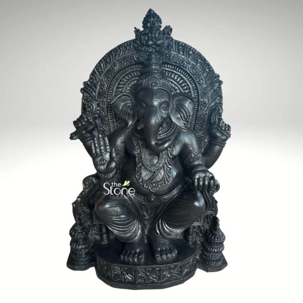 This image features a magnificent 3-foot tall Large Black Stone Lord Ganesha Statue expertly carved from polished black stone. Lord Ganesha is depicted seated in a regal posture, presenting a powerful and auspicious presence against a neutral background. Ganesha is richly adorned with intricate ornaments carved directly into the black stone, including multiple necklaces, armlets, wristlets, and anklets. His large, round belly is prominent, and his trunk curves gracefully to his left, nearing a modak (sweet) held in one of his lower hands. He has four arms; one lower hand is raised in a blessing gesture (Abhaya Mudra), while another holds a broken tusk. His large ears, expressive eyes, and ornate crown with a floral motif add to his divine appearance. The statue is backed by an impressive, intricately carved black stone archway (prabhavali), which features dense traditional scrollwork and decorative patterns, further emphasizing the grandeur of the idol. The entire Ganesha figure sits on a multi-tiered, ornate black stone pedestal, which is also intricately carved with patterns and smaller structural elements, adding to its stability and aesthetic appeal. The polished surface of the black stone reflects light subtly, highlighting the exquisite details of the carvings. This Large Black Stone Lord Ganesha Statue is a grand and beautifully crafted piece, ideal for a temple or a prominent sacred space signifying wisdom and prosperity.