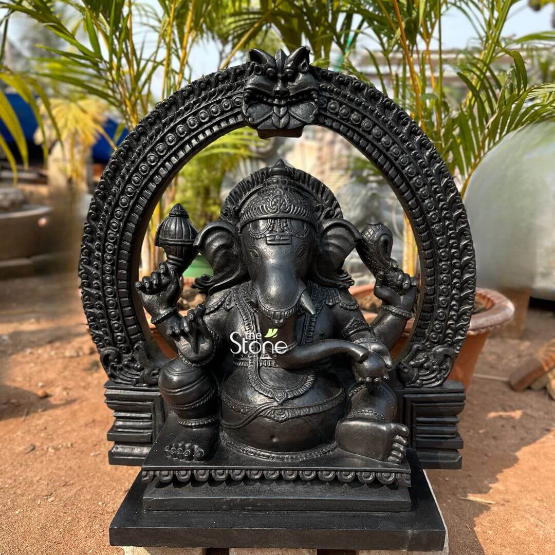 Vighnaharta Ganesh Idol 2ft 3 An exquisitely hand-carved 24-inch Lord Ganapati statue with a decorative arch, masterfully crafted from durable fibre with a refined black granite finish. This 2-foot spiritual masterpiece depicts the deity in a stable and serene seated meditative pose, designed to replicate the majestic, heavy look of natural blackstone while remaining lightweight and weather-resistant. The sculpture features Lord Ganapati framed by a grand, circular Prabhavali (ornate arch), which is embellished with intricate repetitive patterns and a protective Kirtimukha face at its peak, evoking the architectural grandeur of ancient Indian temples. The deity is shown with four arms holding sacred symbols, including a Gada (mace) and a Shankha (conch shell), representing divine power and the removal of obstacles. Every element of this hand-carved black stone-finish Ganesha idol is rendered with precision, from the towering tiered crown (mukut) and layered necklaces to the realistic, fluid folds of his traditional attire. Photographed in an outdoor setting with lush green palm plants in the background, this fibre Ganapati idol serves as a profound spiritual focal point, perfect for a garden landscape, home mandir, or office entrance, inviting peace, stability, and auspicious energy into any environment.