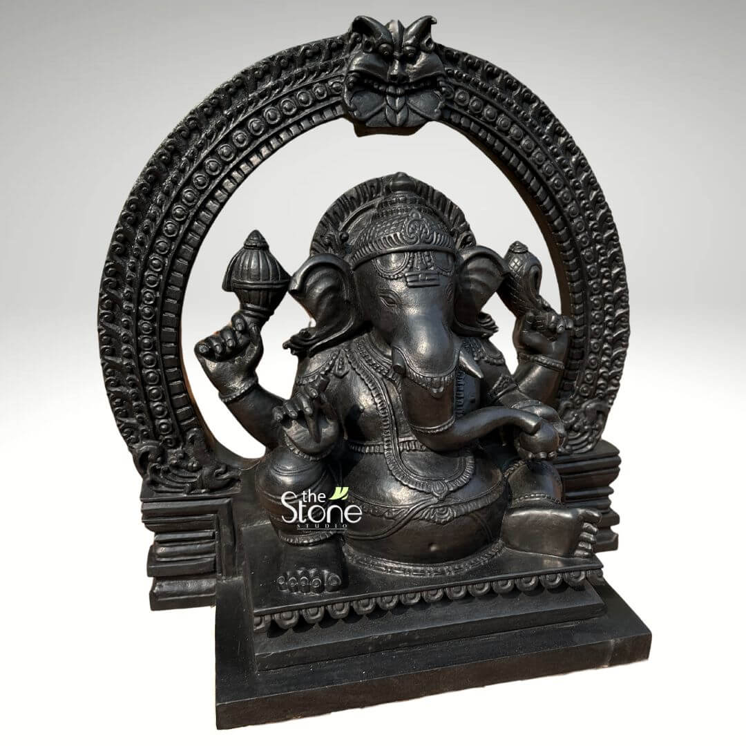 Vighnaharta Ganesh Idol 2ft 7 An exquisitely hand-carved 24-inch Lord Ganesha black granite-finish statue, featuring a majestic Prabhavali (ornate arch). This 2-foot fibre statue is masterfully designed to replicate the heavy, timeless look of natural blackstone, making it a durable and lightweight alternative for modern spiritual spaces. The sculpture depicts Lord Ganesha in a stable and serene seated meditative pose, framed by a grand circular archway. The arch is embellished with intricate repetitive patterns and a protective Kirtimukha face at the peak, evoking the architectural grandeur of ancient Indian temples. The deity is shown with four arms holding sacred symbols, including a Gada (mace) and a Shankha (conch shell), while his lower right hand is raised in the Abhaya Mudra of blessing. Every detail, from the towering tiered crown (mukut) and layered necklaces to the realistic, fluid folds of his traditional attire, is rendered with exceptional precision. The statue rests on a solid, multi-tiered rectangular pedestal featuring a delicate relief border. This black stone-finish Ganesha idol serves as a profound spiritual focal point, perfect for a home mandir, office foyer, or meditation sanctuary, designed to invite peace, stability, and auspicious energy into any living environment.