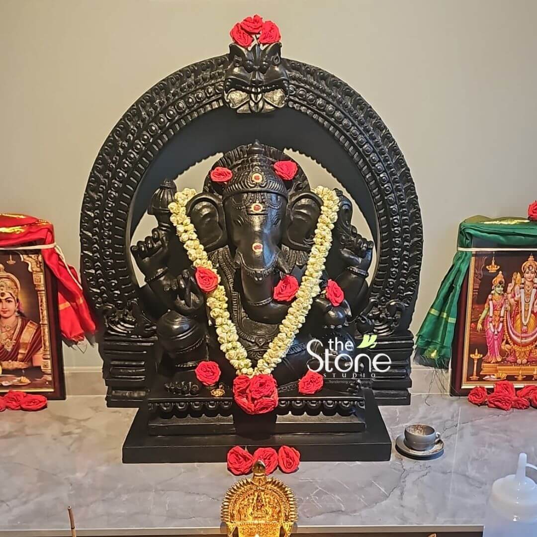 Vighnaharta Ganesh Idol 2ft 6 This image features a majestic 2-foot tall Lord Ganesha Statue carved from polished blackstone, prominently displayed on an altar, possibly in a home or temple in Singapore. Lord Ganesha is seated in a traditional posture, adorned with fresh floral decorations. The blackstone statue of Ganesha is the central focus, emanating a powerful presence. He is adorned with a beautiful garland of white flowers, intertwined with vibrant red roses, draped around his neck. Additional red roses are placed on his head, ears, and hands, as well as scattered on the blackstone base in front of him. His eyes are prominent, and a decorative element, possibly a bindi, is placed on his forehead. He holds his traditional attributes in his multiple arms. The Ganesha statue is framed by an impressive blackstone archway (prabhavali), which is intricately carved with traditional motifs and textured details. At the top of the arch, a fierce Kirtimukha motif is visible. The entire setup rests on a dark, patterned marble surface. Flanking the Ganesha statue are two framed devotional pictures, draped with red and green cloths respectively, adding to the sacred ambiance. In front of the statue, on the marble surface, are a few offerings: a small, intricately designed golden Ganesha murti, a tiny cup, and possibly an incense holder or other ritualistic items. The overall presentation is one of profound devotion, highlighting this elegant blackstone Ganesha statue as a revered deity.