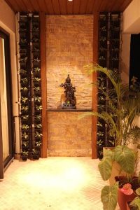 A serene spiritual nook in a modern Bangalore home featuring a 3-foot tall Lord Krishna statue hand-carved from natural blackstone. The deity is depicted in a classic devotional posture, gracefully playing the flute. The statue is positioned on a dark floating shelf, which serves as a centerpiece against a textured, warm-toned stacked stone feature wall. The entire vertical installation is flanked by two columns of a vertical garden, with small black planters housing lush green foliage that adds a touch of nature to the indoor space. Soft, warm overhead lighting casts a gentle glow on the Krishna idol, highlighting its intricate carvings and creating a peaceful, meditative atmosphere. In the foreground, the edges of large tropical plants and a patterned tile floor complete the elegant foyer or balcony decor, showcasing how traditional spiritual art can be integrated into contemporary urban living in Bangalore.