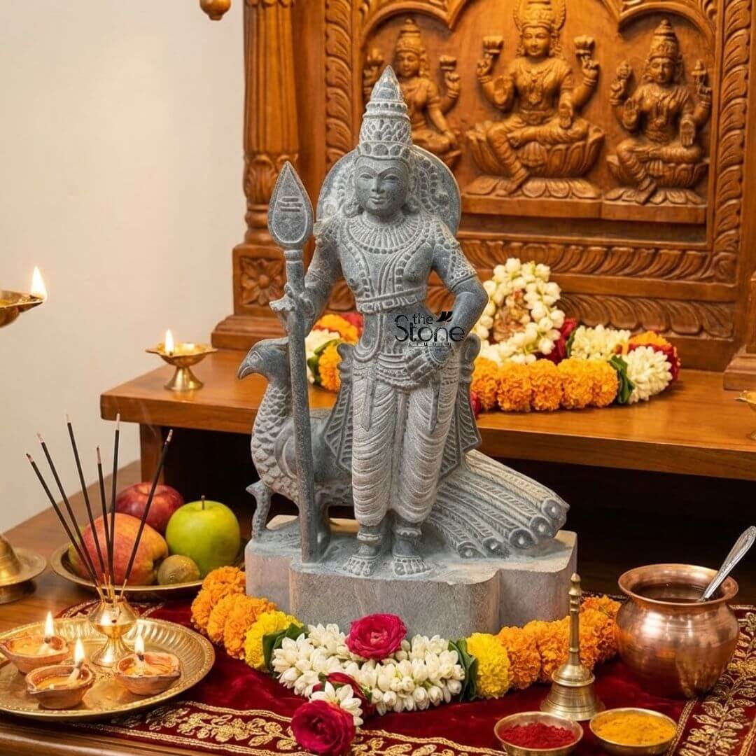 Lord Skanda Murugan Stone Statue 15" 5 A serene and devotional home altar featuring an exquisitely hand-carved 15-inch Lord Murugan stone idol with Vel and peacock. Masterfully sculpted from natural blackstone with a rustic grey finish, the deity stands in a majestic posture, holding his signature sacred spear (Vel) in his right hand, representing divine wisdom and protection. His faithful vehicle, the peacock, is delicately carved behind him with its tail feathers fanning out in detailed relief. The Murugan stone statue is artfully positioned on a wooden altar draped in a rich red velvet cloth with golden embroidery. It is surrounded by an auspicious arrangement of fresh offerings, including vibrant garlands of yellow and orange marigolds, white jasmine strings, and red roses. The devotional setting is further enhanced by two lit brass oil lamps (diyas), burning incense sticks on a gold-toned tray, a brass ritual bell, and copper vessels containing kumkum and turmeric. In the background, a beautifully carved dark wooden mandir panel depicts traditional Indian deities, illustrating how this 15-inch blackstone Lord Murugan idol serves as a profound spiritual centerpiece for prayer and meditation in a traditional home sanctuary.