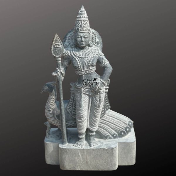 An exquisitely hand-carved 15-inch Lord Skanda Murugan stone statue, masterfully sculpted from premium natural blackstone to serve as a powerful spiritual centerpiece. The deity, also known as Kartikeya or Subramanya, is depicted in a majestic standing posture, holding his iconic Vel (sacred spear) in his right hand, which symbolizes divine wisdom and the destruction of ignorance. Lord Murugan is adorned with an intricately detailed tiered crown (mukut), layered necklaces, armlets, and traditional ornaments that showcase the depth of Indian stone craftsmanship. Standing faithfully behind the Lord is his divine vehicle, the peacock (vahana), with its tail feathers delicately rendered in relief. The blackstone Murugan idol features a rustic, weathered grey finish that highlights the fine textures of the stone masonry. Resting on a sturdy, multi-lobed stone pedestal, this 15-inch spiritual sculpture is an ideal size for a home mandir, personal altar, or meditation corner. It captures the serene yet strong essence of the deity, inviting an atmosphere of protection, victory, and peace into any sacred living space.