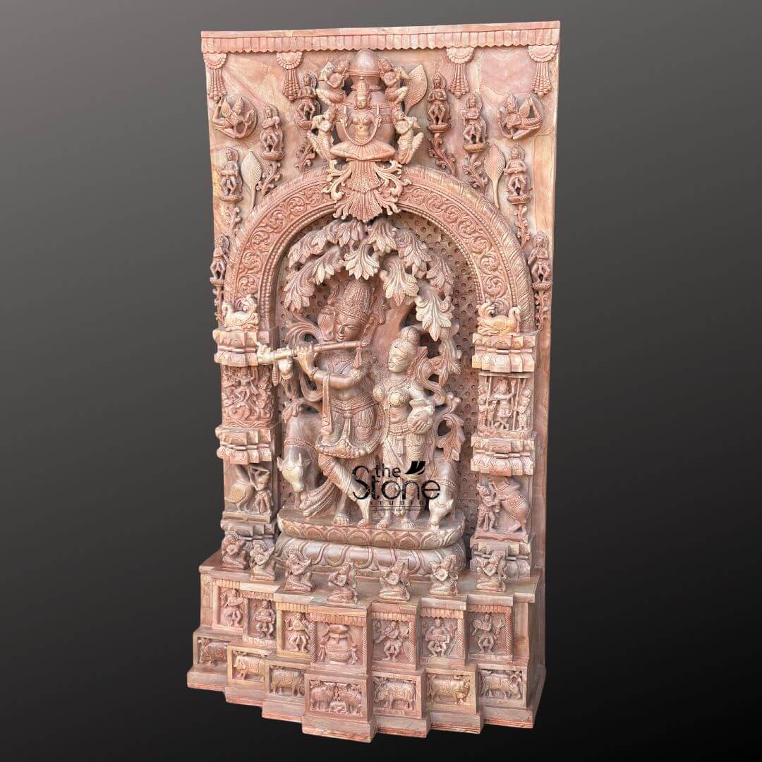 Pink Stone Radha Krishna Murti for Home 4ft 2 This image showcases a magnificent 4-foot tall Luxury Radha Krishna Idol meticulously carved from natural pink stone, presented as a complete sculptural panel. The central and most prominent feature is the depiction of Lord Krishna playing his flute, standing gracefully with crossed legs beneath a intricately detailed tree. Beside him, Radha stands in an attentive and loving posture, both figures adorned with traditional garments and jewelry, all exquisitely rendered in the soft pink stone. The main scene of Radha and Krishna is framed within an elaborate, arch-shaped niche, which is itself surrounded by a rich tapestry of carvings. Above the arch, at the very top of the panel, a grander deity figure, possibly Vishnu or another celestial being, is enthroned, flanked by smaller attendant figures and intricate architectural motifs. The entire panel is a testament to skilled craftsmanship, with every inch filled with minute details including flora, fauna, mythological creatures, and numerous smaller divine figures or devotees. These surrounding carvings add layers of narrative and symbolic depth, enhancing the central theme. The lower section of the panel features multiple tiers of carved friezes and smaller alcoves, each containing detailed sculptures, further contributing to the grandeur and 'luxury' aspect of the piece. The natural pink hue of the stone adds a unique warmth and aesthetic appeal, making this an ideal statement piece for a discerning collector or a prominent feature in a devotional space. The entire composition emphasizes the divine love and beauty of Radha and Krishna in an opulent and timeless manner.