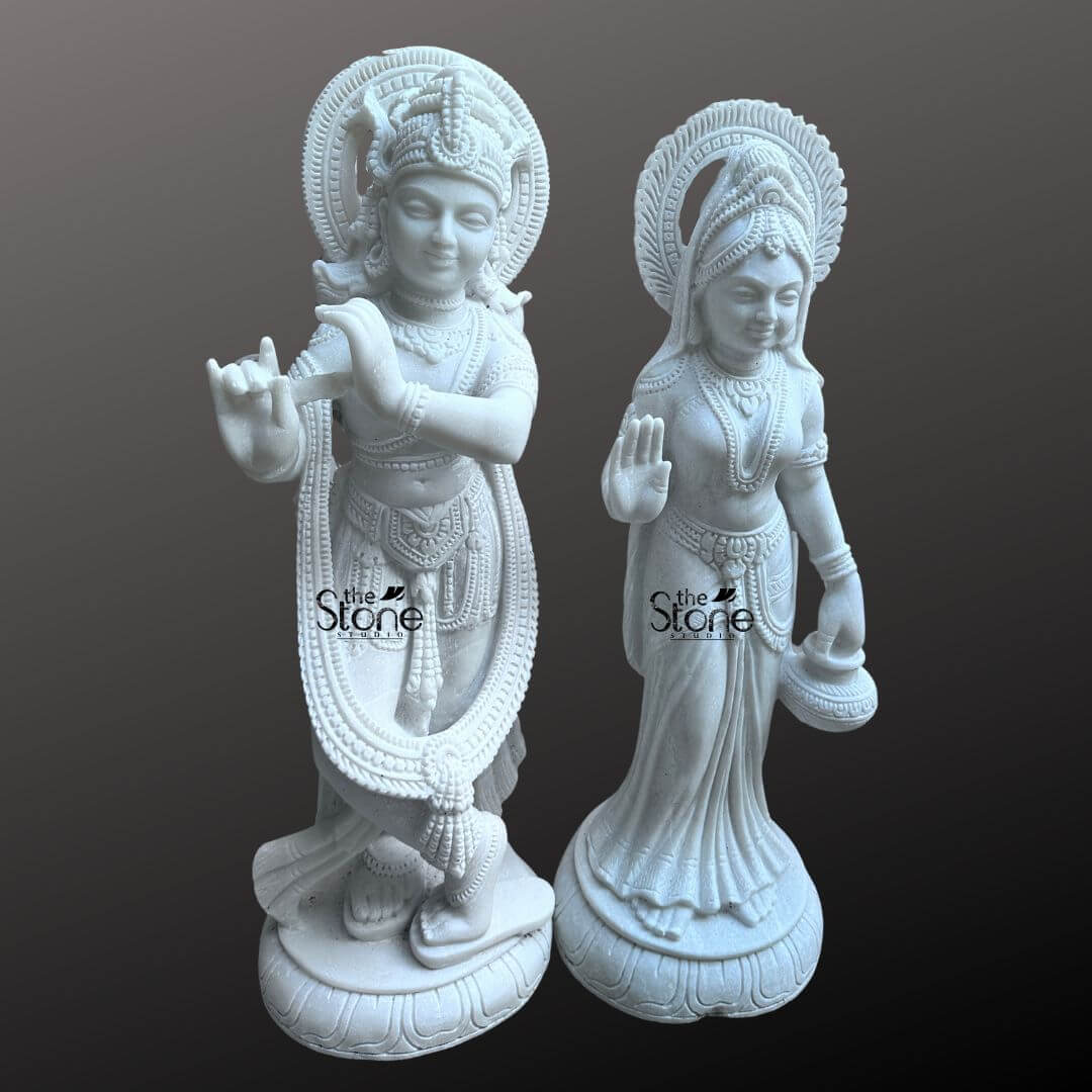 Radha Krishna Marble Stone Statue 3ft 8 This image presents a stunning Marble Stone Radha Krishna Sculpture featuring a pair of 3-foot tall white marble statues of Radha and Krishna, expertly carved and displayed against a dark, neutral background. The pristine white marble highlights the intricate details and graceful forms of the deities. On the left, Lord Krishna stands in his iconic Tribhanga posture, playing his flute. He is richly adorned with finely sculpted jewelry, a detailed crown, and flowing garments, all rendered with precision in the white marble. A long garland also encircles his form. A subtle circular halo is carved behind his head. On the right Radha stands gracefully, her right hand raised in a gesture of blessing and her left hand holding a small pot or offering. She is also beautifully adorned with a crown, necklaces, bracelets, and a flowing sari, all intricately carved from the same white marble. A similar circular halo is behind her head. Both statues stand on individual, ornate lotus pedestals, which are integrated into the overall marble carving. The smooth polished surface of the "white marble statue" reflects light softly, emphasizing the delicate features and the divine expressions of Radha and Krishna. This Marble Stone Radha Krishna Sculpture is a beautiful embodiment of sacred love and traditional Indian artistry, ideal for a temple or a prominent place in a home.