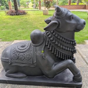 A majestic fiber statue of Lord Nandi, the sacred bull and loyal vehicle of Lord Shiva, is beautifully showcased as a spiritual centerpiece in a lush Coimbatore garden. Masterfully crafted from durable and lightweight fiber material, the sculpture features a refined matte black finish that masterfully replicates the timeless look of natural black stone. The Nandi idol is depicted in a traditional recumbent posture, resting on a solid integrated rectangular base atop a stone patio. The sculpture displays exceptional craftsmanship with highly intricate detailing, including multiple layers of carved bell necklaces and beaded garlands around its neck. A richly detailed caparison (decorative blanket) featuring rhythmic floral relief patterns and a large central mandala is draped across its back and prominent hump. Set against a backdrop of a manicured green lawn and tropical trees, this weather-resistant fiber Nandi sculpture illustrates how traditional Vedic iconography can be seamlessly integrated into modern outdoor garden decor in Coimbatore. It serves as an auspicious guardian figure, inviting strength, devotion, and a sense of sacred tranquility into the home courtyard or temple landscape.