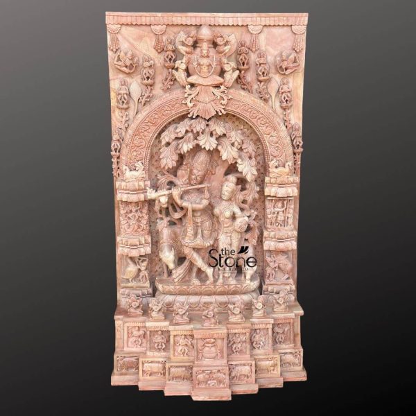 A magnificent 4-foot tall pink stone carving depicts Radha and Krishna as the central figures within a highly ornate, rectangular panel. The exquisite craftsmanship is evident in the intricate details carved from the natural pink stone, which gives the artwork a warm and inviting hue. At the heart of the panel, Lord Krishna is shown playing his flute under a beautifully sculpted tree, with his legs gracefully crossed. Beside him, Radha stands attentively, captivated by his music, her form also delicately carved with traditional attire and jewelry. The figures are framed by an elaborate arched niche, adorned with further intricate patterns and foliage, suggesting a lush Vrindavan setting. Above the main Radha Krishna scene, at the very top of the panel, a larger deity figure, possibly Vishnu or a celestial being, is enthroned, surrounded by smaller divine or semi-divine figures. This upper section is connected to the central arch by rich, detailed carvings. The sides of the panel are also filled with numerous smaller niches and figures, including devotees, animals, and mythological creatures, all meticulously sculpted in the same pink stone, adding depth and narrative to the entire composition. The base of the panel features a series of additional carved friezes and smaller devotional scenes, suggesting stories or aspects of the divine pair's lives. The sheer density of the carving and the warm pink tone of the stone make this a striking piece, perfectly suited as a prominent Radha Krishna Murti for Home, radiating devotion and artistic splendor.