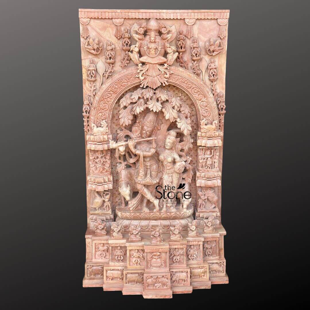 Pink Stone Radha Krishna Murti for Home 4ft 1 A magnificent 4-foot tall pink stone carving depicts Radha and Krishna as the central figures within a highly ornate, rectangular panel. The exquisite craftsmanship is evident in the intricate details carved from the natural pink stone, which gives the artwork a warm and inviting hue. At the heart of the panel, Lord Krishna is shown playing his flute under a beautifully sculpted tree, with his legs gracefully crossed. Beside him, Radha stands attentively, captivated by his music, her form also delicately carved with traditional attire and jewelry. The figures are framed by an elaborate arched niche, adorned with further intricate patterns and foliage, suggesting a lush Vrindavan setting. Above the main Radha Krishna scene, at the very top of the panel, a larger deity figure, possibly Vishnu or a celestial being, is enthroned, surrounded by smaller divine or semi-divine figures. This upper section is connected to the central arch by rich, detailed carvings. The sides of the panel are also filled with numerous smaller niches and figures, including devotees, animals, and mythological creatures, all meticulously sculpted in the same pink stone, adding depth and narrative to the entire composition. The base of the panel features a series of additional carved friezes and smaller devotional scenes, suggesting stories or aspects of the divine pair's lives. The sheer density of the carving and the warm pink tone of the stone make this a striking piece, perfectly suited as a prominent Radha Krishna Murti for Home, radiating devotion and artistic splendor.
