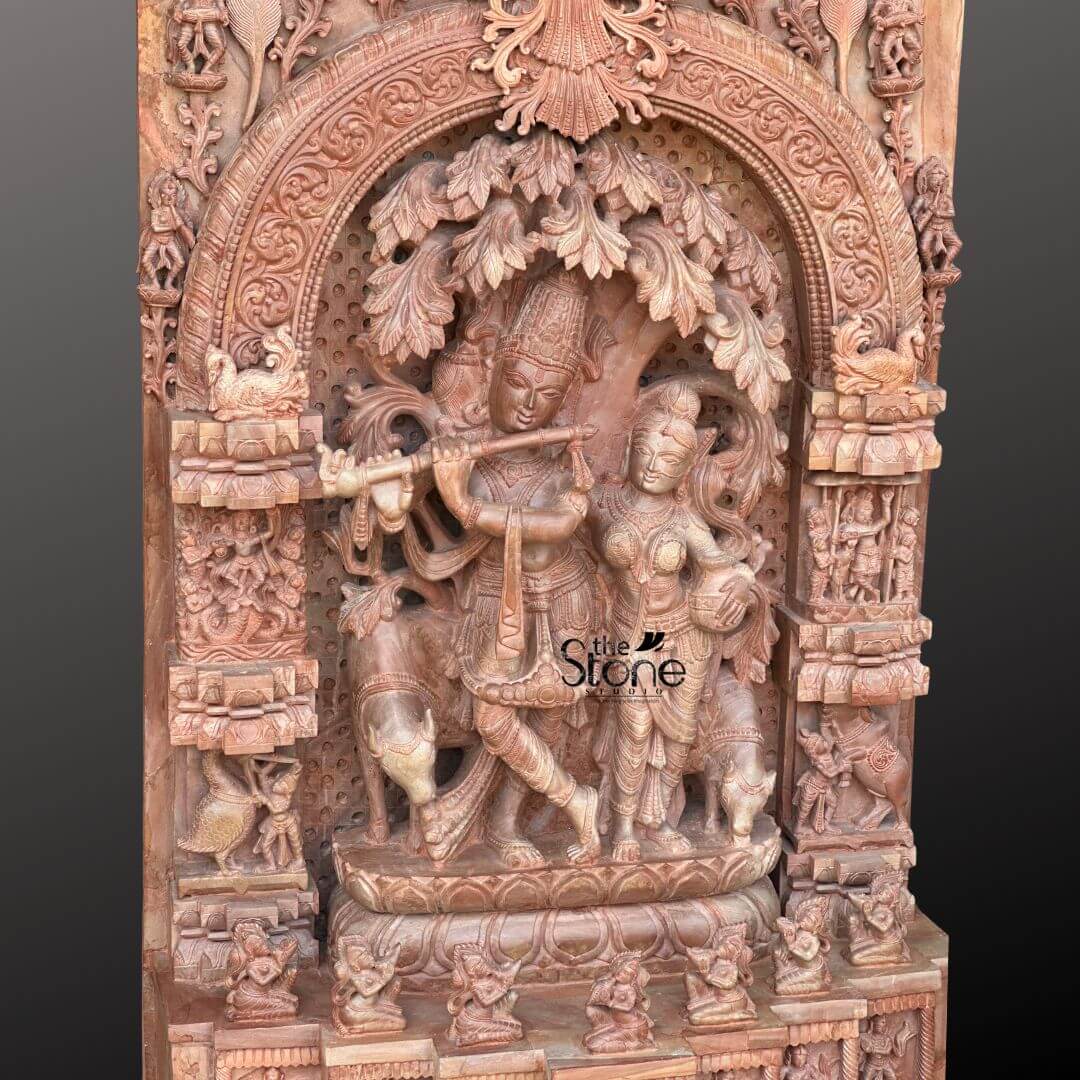 Pink Stone Radha Krishna Murti for Home 4ft 4 This is a close-up, highly detailed view of a 4-foot tall Pink Stone Radha Krishna Murti with Detailed Carvings. The central focus is the exquisitely sculpted depiction of Lord Krishna playing his flute, standing with one leg crossed, under a beautifully rendered tree whose leaves and branches form an intricate canopy behind him. Beside him, Radha stands attentively, her form also richly detailed with traditional attire, jewelry, and a gentle expression, all carved from the warm pink stone. The main Radha Krishna scene is framed within a deeply carved, ornate archway that forms the centerpiece of the larger panel. The carving density is exceptional, showcasing incredibly fine details that highlight the craftsmanship. To Krishna's left, a cow is partially visible, adding to the idyllic Vrindavan setting. The surrounding areas of the panel are equally laden with detailed carvings. Along the sides of the arch, vertical columns are filled with numerous smaller figures in niches, depicting devotees, celestial beings, and various mythological elements. The top section of the arch features intricate floral patterns and divine representations. Below the main figures, the pedestal is adorned with multiple tiers of smaller sculptures and decorative motifs, creating a sense of grandeur and historical depth. The natural pink hue of the stone further enhances the intricate textures and shadows, making every detail stand out and emphasizing the luxurious and artistic quality of this devotional piece.
