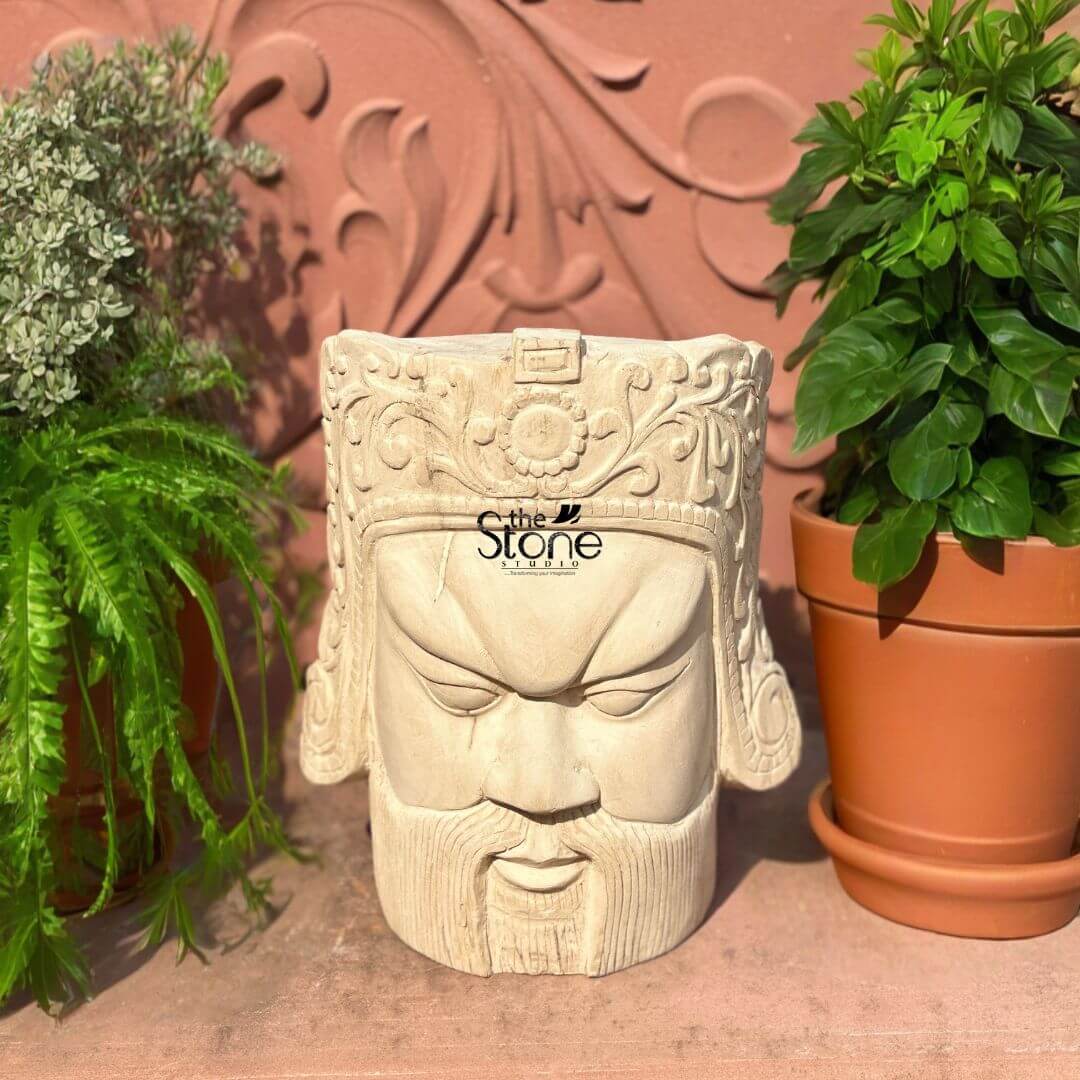 Royal Head Sculpture 12" 4 This image showcases a Premium Hand Carved Wooden Mask Style Sculpture a 12-inch male head carved from light-colored wood presented in an outdoor or garden setting. The sculpture has a commanding and somewhat fierce expression, reminiscent of ancient warrior masks or guardian figures. The face features strong, angular characteristics: deeply furrowed brows, intense, narrow eyes that gaze downward, a straight nose, and a firmly closed mouth adorned with a full, sculpted beard and mustache. The carving creates pronounced shadows, enhancing the dramatic and powerful expression. Crowning the head is an elaborate headdress, richly detailed with intricate floral patterns, swirling motifs, and a central circular emblem. The precision of the hand carved details is remarkable, with every element meticulously shaped from the wood. The natural grain and subtle imperfections of the wood are visible throughout the bust, adding to its organic texture and handcrafted appeal. The sculpture is placed on a reddish-brown, possibly terracotta, surface, with two potted green plants flanking it. In the background, a decorative wall with ornate reddish-brown carvings adds to the aesthetic, providing a natural and earthy context for this unique wooden head sculpture. The overall presentation highlights the sculpture's artistic merit and its potential as a distinctive decorative piece.