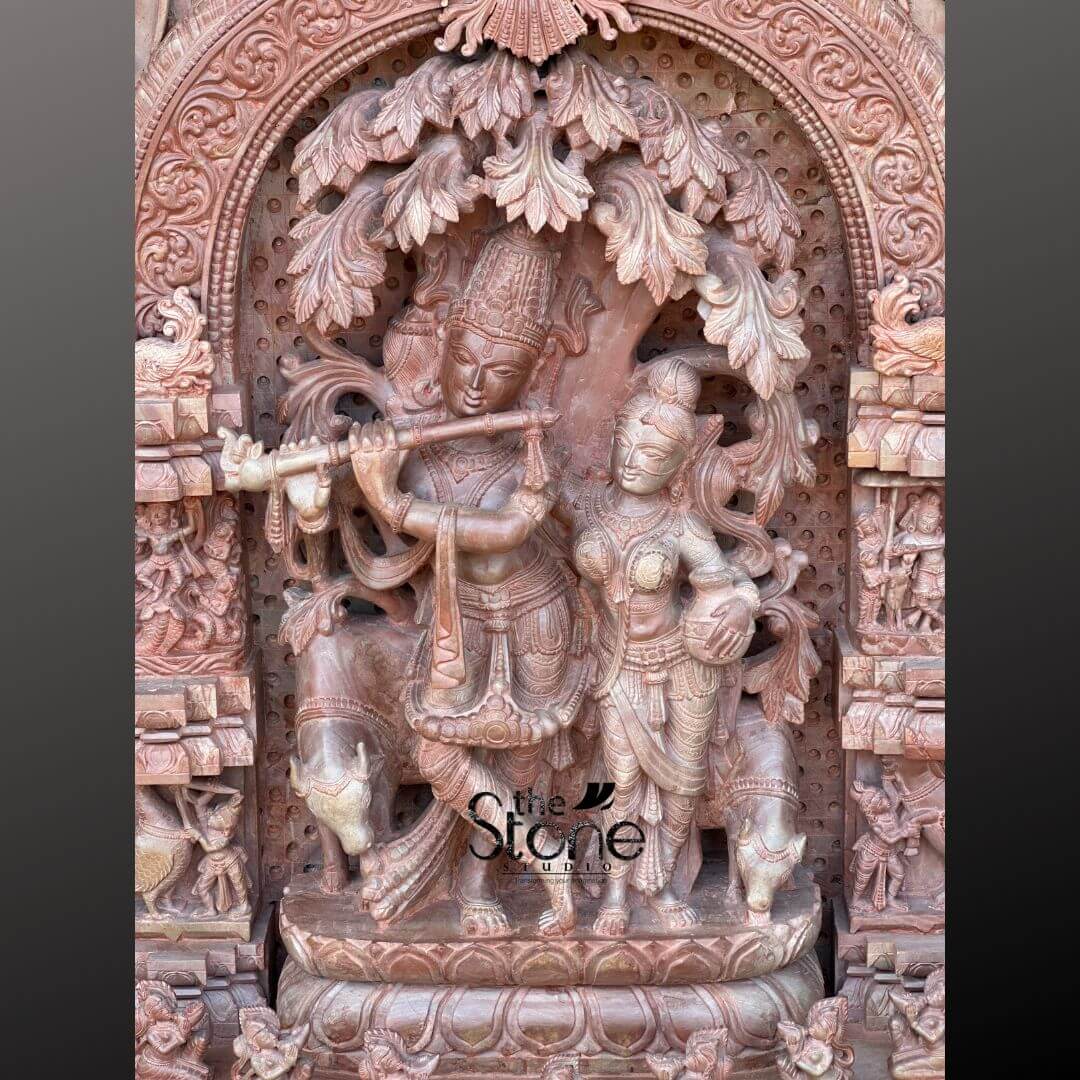 Pink Stone Radha Krishna Murti for Home 4ft 6 This close-up image presents a magnificent Radha Krishna Mandir Panel carved from premium pink stone, serving as a prominent feature for a temple or home shrine. The 4-foot tall sculpture is incredibly detailed, with the central figures of Lord Krishna playing his flute and Radha standing gracefully beside him, both rendered with exceptional artistry. Krishna, with his left leg crossed over his right, holds the flute to his lips, while Radha, holding a pot or vessel, gazes towards him. They stand beneath a beautifully carved tree, its extensive foliage and branches creating a lush, organic backdrop. The intricate details extend to their crowns, jewelry, and flowing garments, all sculpted with remarkable precision from the natural pink stone, which highlights the contours and textures. To the left of Krishna, a partially visible cow adds to the pastoral charm of the scene. The entire central tableau is framed by an elaborate, deeply carved archway, itself a work of art with floral motifs, divine figures, and architectural elements. The columns flanking the arch are filled with numerous smaller devotional figures and narrative scenes, meticulously chiseled into the stone. Below the main figures, the multi-tiered base is also adorned with intricate carvings, including smaller sculptures of devotees and animals, adding to the panel's grandeur and sacred storytelling. The use of premium pink stone gives the entire 4-foot Radha Krishna Statue a warm, inviting, and luxurious feel, truly embodying the essence of a divine mandir panel.