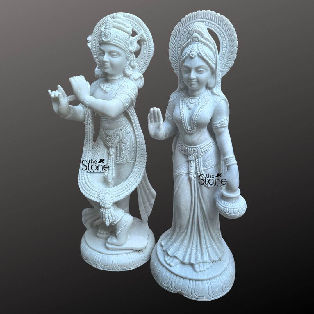 Radha Krishna Marble Stone Statue 3ft 7 This image presents a stunning Radha Krishna Marble Sculpture with Carvings featuring a pair of 3-foot tall white marble statues of Radha and Krishna. Expertly carved the deities are displayed against a dark neutral background allowing the pristine white marble and intricate details to stand out. On the left, Lord Krishna stands in his iconic Tribhanga posture, playing his flute. He is richly adorned with finely sculpted jewelry, a detailed crown and flowing garments, all rendered with precision in the white marble. A long garland also encircles his form. A subtle circular halo with carvings is behind his head. On the right, Radha stands gracefully her right hand raised in a gesture of blessing and her left hand holding a small pot or offering. She is also beautifully adorned with a crown, necklaces, bracelets, and a flowing sari, all intricately carved from the same white marble. A similar circular halo with fine carvings is behind her head. Both statues stand on individual, ornate lotus pedestals, which are integrated into the overall marble carving and feature additional carved details. The smooth, polished surface of the white marble statue reflects light softly, emphasizing the delicate features, the intricate carvings, and the divine expressions of Radha and Krishna. This Radha Krishna Marble Sculpture is a beautiful embodiment of sacred love and traditional Indian artistry, ideal for a temple or a prominent place in a home.