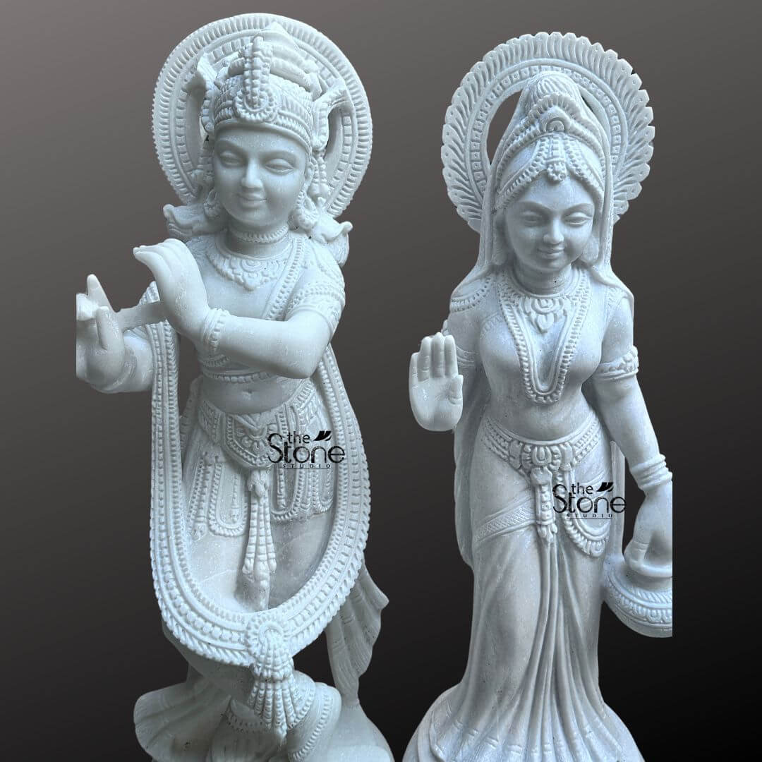 Radha Krishna Marble Stone Statue 3ft 6 This image presents a stunning Radha Krishna Marble Statue for Home featuring a pair of 3-foot tall white marble statues of Radha and Krishna. Expertly carved, the deities are displayed against a dark, neutral background, allowing the pristine white marble and intricate details to stand out. On the left, Lord Krishna stands in his iconic Tribhanga posture, playing his flute. He is richly adorned with finely sculpted jewelry, a detailed crown, and flowing garments, all rendered with precision in the white marble. A long garland also encircles his form. A subtle circular halo with delicate carvings is behind his head. On the right Radha stands gracefully her right hand raised in a gesture of blessing and her left hand holding a small pot or offering. She is also beautifully adorned with a crown, necklaces, bracelets, and a flowing sari, all intricately carved from the same white marble. A similar circular halo with fine carvings is behind her head. Both statues stand on individual, ornate lotus pedestals, which are integrated into the overall marble carving and feature additional carved details. The smooth, polished surface of the white marble statue reflects light softly, emphasizing the delicate features, the intricate carvings, and the divine expressions of Radha and Krishna. This Radha Krishna Marble Statue is a beautiful embodiment of sacred love and traditional Indian artistry, ideal for a prominent place in a Home or a private shrine.