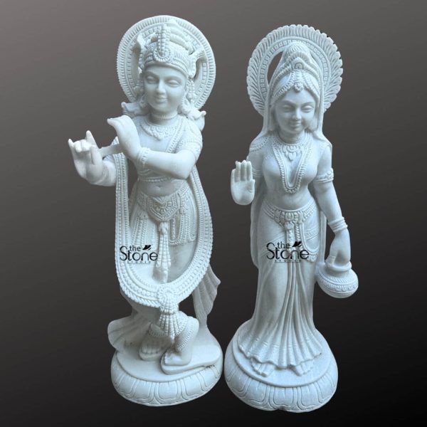 This image presents a stunning Radha Krishna Marble Stone Statue featuring a pair of 3-foot tall white marble statues of Radha and Krishna. Expertly carved the deities are displayed against a dark neutral background allowing the pristine white marble and intricate details to stand out. On the left Lord Krishna stands in his iconic Tribhanga posture holding and playing his flute. He is richly adorned with finely sculpted jewelry a detailed crown with a peacock feather and flowing garments all rendered with exceptional precision in the white marble. A long garland also encircles his form. A prominent intricately carved circular halo is behind his head signifying his divine aura. On the right Radha stands gracefully her right hand raised in a gesture of blessing and her left hand holding a small pot or offering. She is also beautifully adorned with a crown, necklaces, bracelets, and a flowing sari all intricately carved from the same white marble. A similar finely carved circular halo is behind her head. Both statues stand on individual ornate lotus pedestals which are integrated into the overall marble carving and feature additional carved details. The smooth polished surface of the white marble statue reflects light softly emphasizing the delicate features the extensive carvings on their attire and jewelry and the divine expressions of Radha and Krishna. This Radha Krishna Marble Stone Statue is a beautiful embodiment of sacred love and traditional Indian artistry ideal for a temple or a prominent place in a home.